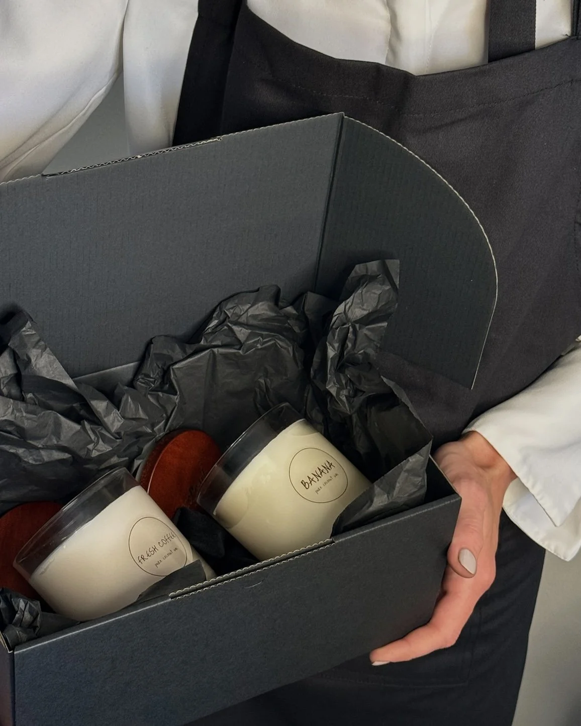 A person holding a black box containing two candles labeled 'FRESH COCONUT' and 'BANANA' in glass jars, with crumpled black tissue paper inside the box.
