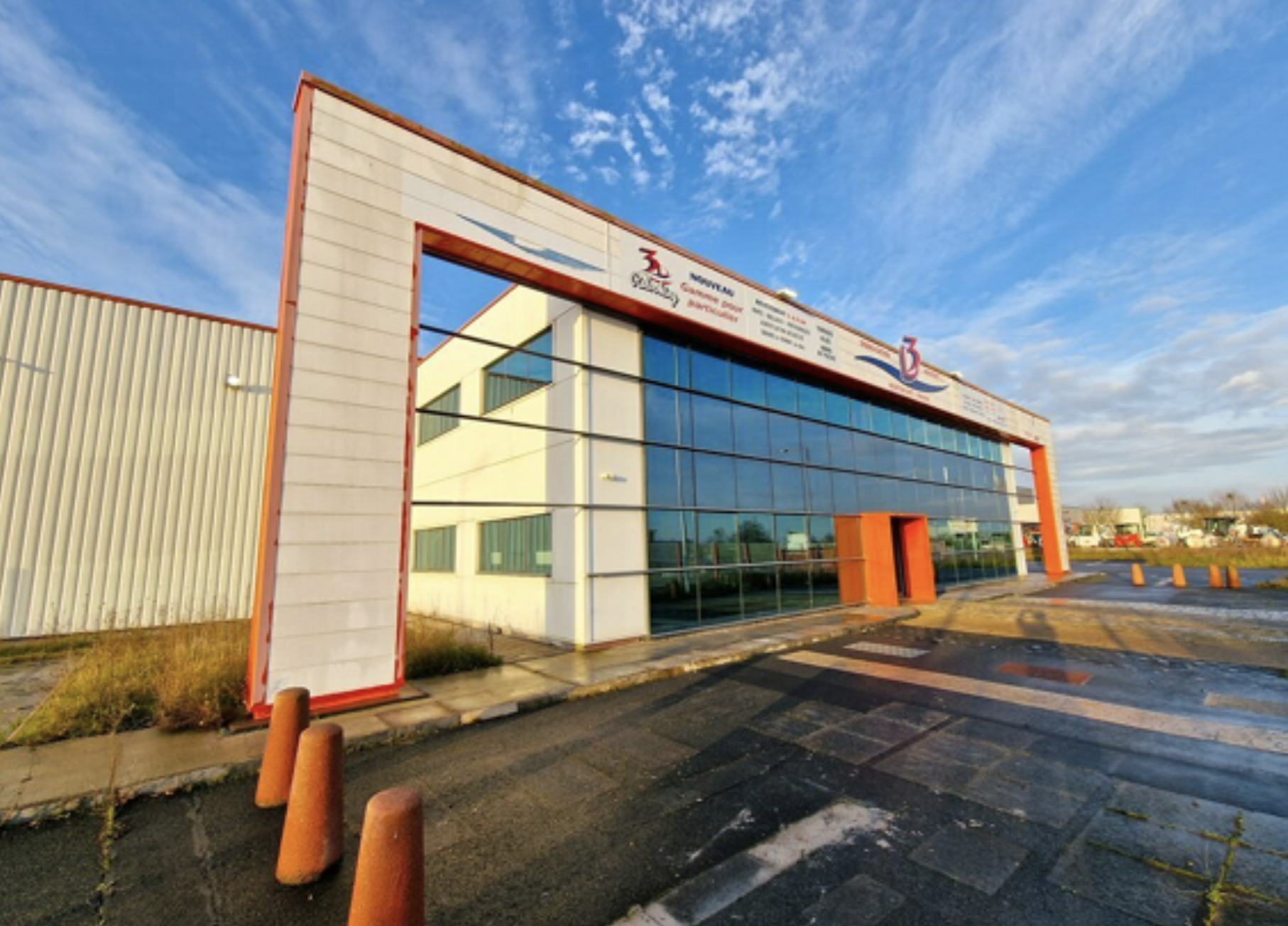 Bâtiment moderne avec grande façade en verre, panneaux d'affichage en haut, et un parking devant, sous un ciel bleu avec quelques nuages.