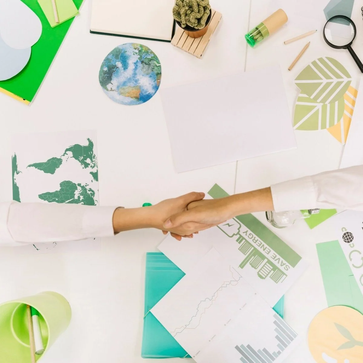 Deux personnes se serrant la main sur un bureau avec des documents, graphiques écologiques, une plante en pot, et des fournitures de bureau.