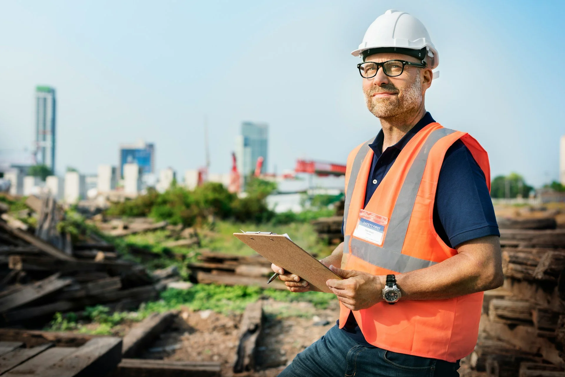 Man wearing safety vest and hard hat holding a clipboard at a construction site with city buildings in the background.
