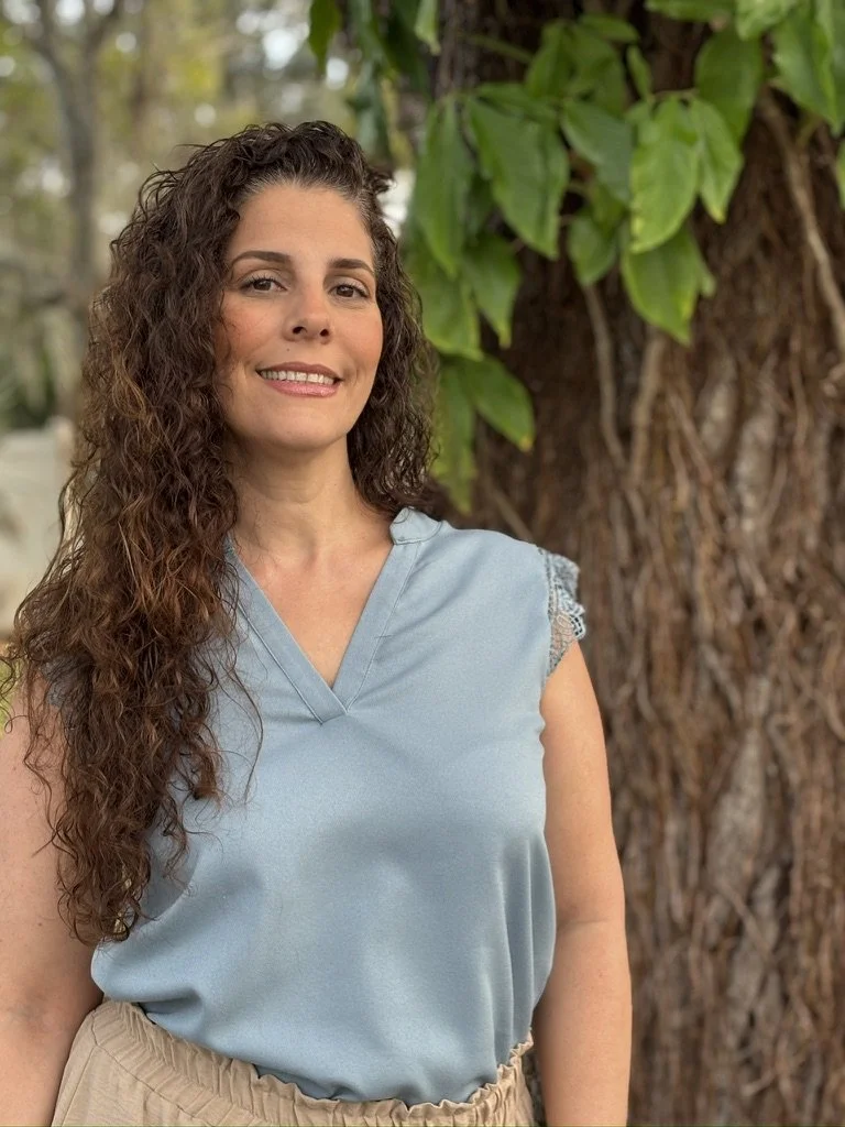 A woman with long curly brown hair wearing a light blue sleeveless top, standing outdoors near a large tree with green leaves.