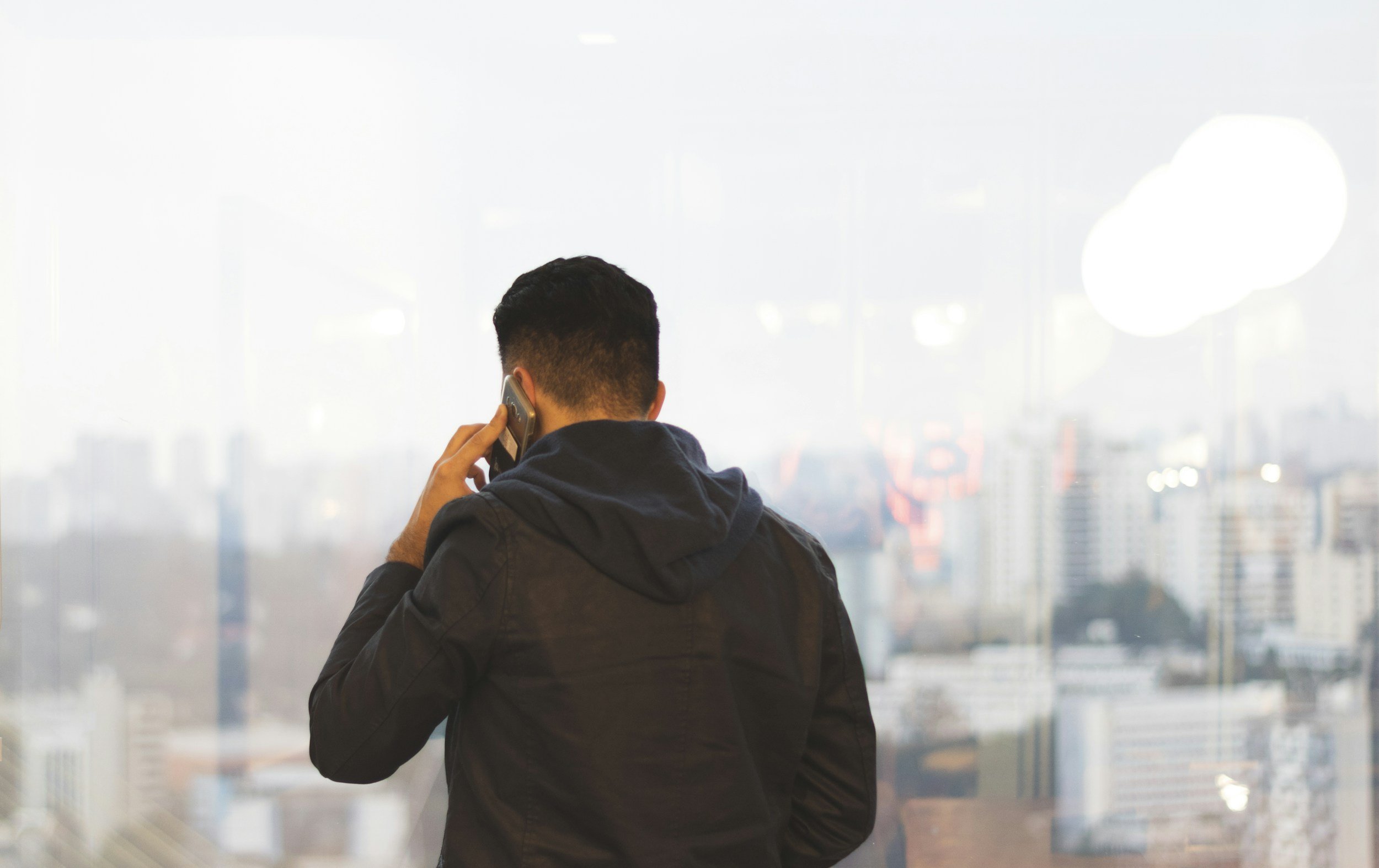 A man with short dark hair, wearing a black jacket, is talking on a cell phone while facing a large window with a blurred city skyline in the background.