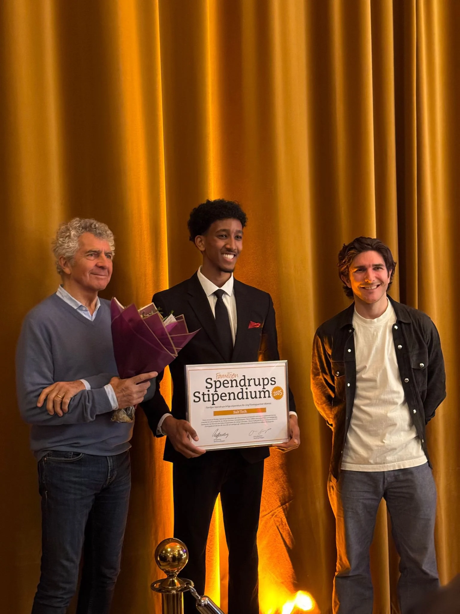 Three men standing on stage with a gold curtain backdrop. The man in the center holds a certificate and a bouquet of flowers, smiling. The man on the left is wearing a gray sweater and jeans, and the man on the right is dressed casually in a black jacket and gray pants, all smiling.