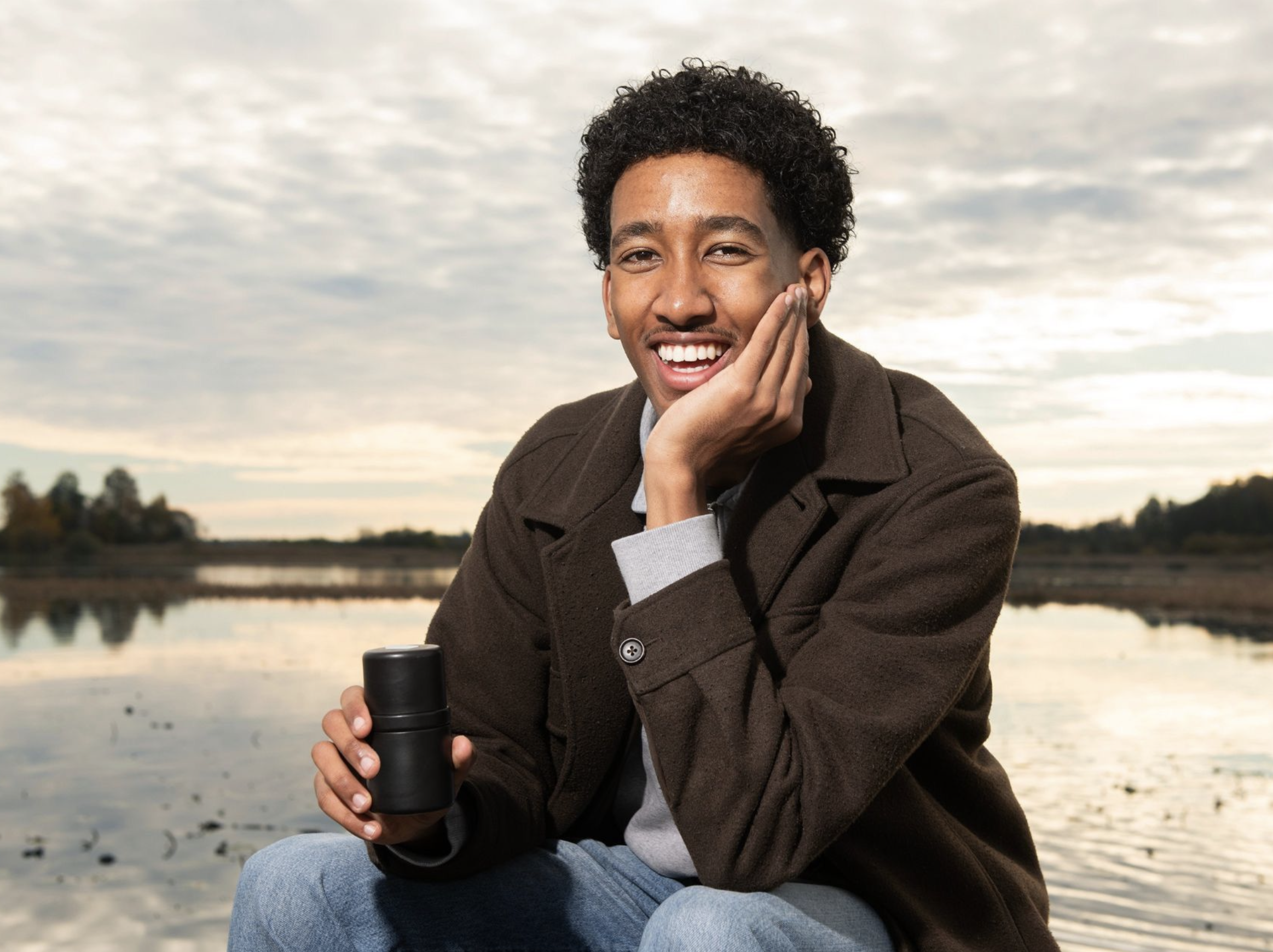 A young man with curly hair smiling and holding a black camera while sitting outdoors near a body of water during sunset.