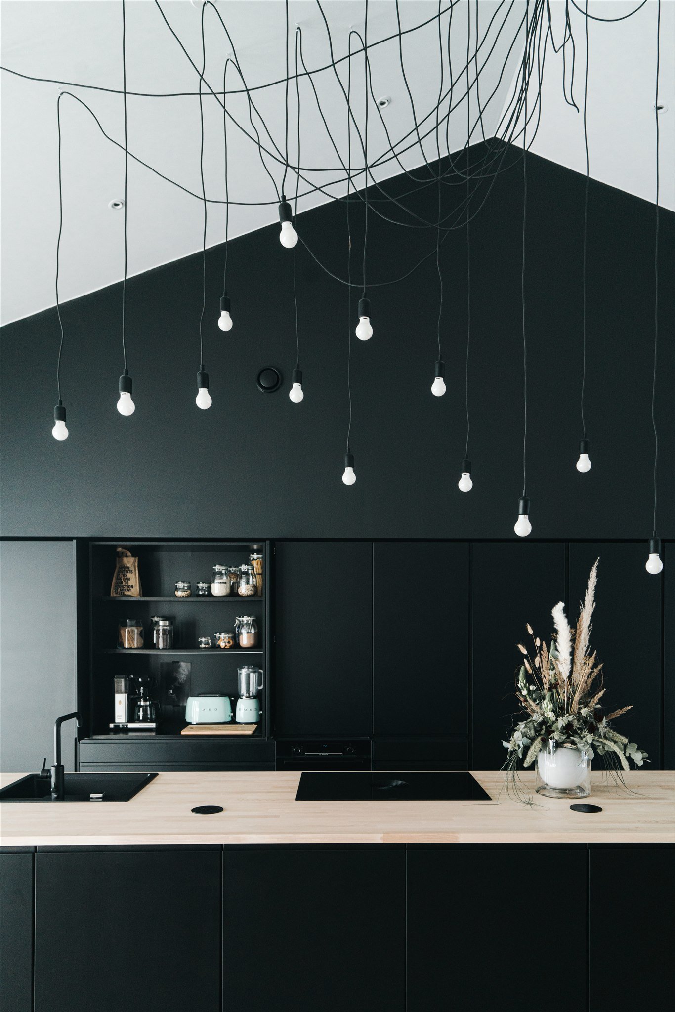 Tyylikäs keittiö, jossa musta kaapisto ja puupöytä, ripaus kuivakukkia maljakossa ja kattovaloja.