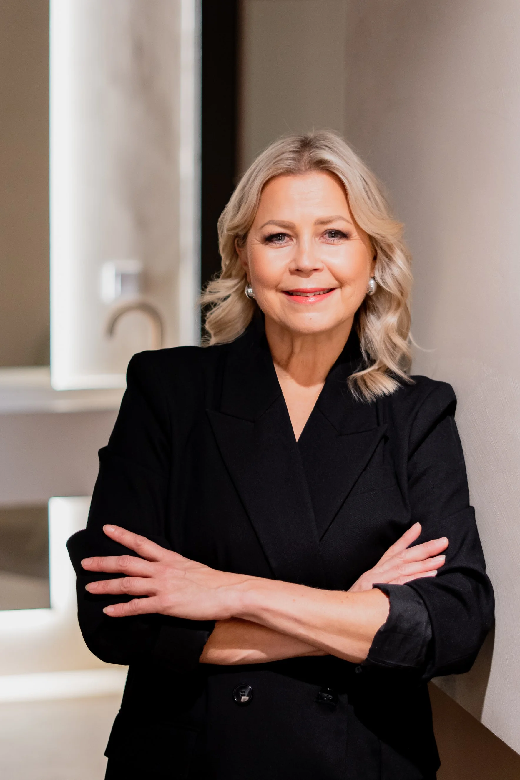 Nainen hymyilee ja poseeraa kädet ristissä, pukeutuneena mustaan pukuun, sisätiloissa.