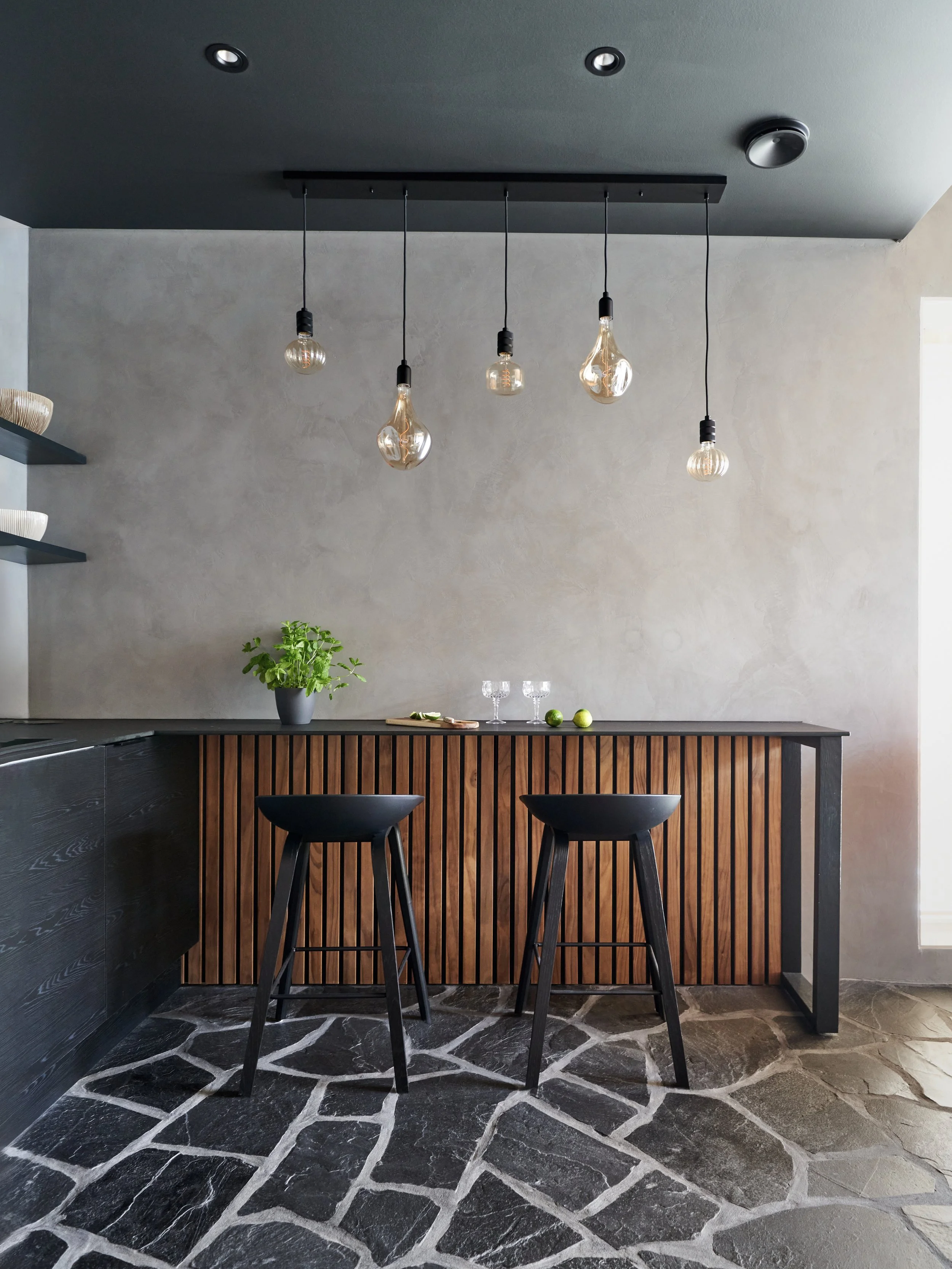 Modernin keittiön sisustus, jossa rosoiset mustat ja kivi lattiat, mustat tuolit ja puurivinen allas, jossa on vihreä kasvi ja laseja pöydällä, taustalla harmaa seinä ja kattoon ripustettu koristeellinen valaisinjärjestelmä.