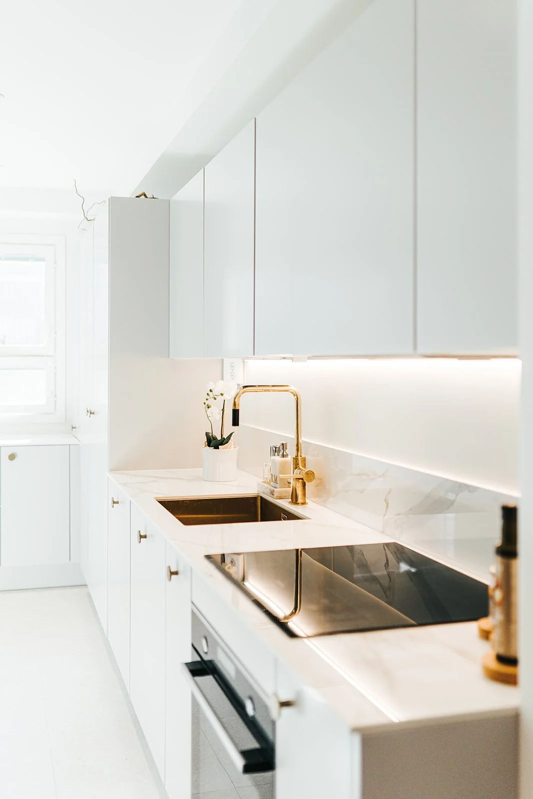 Valkoinen keittiö, jossa kullansävyiset vesihanat ja tekevät työtasot, pieni kasvi ja kiviä sisältävä kaakelo.