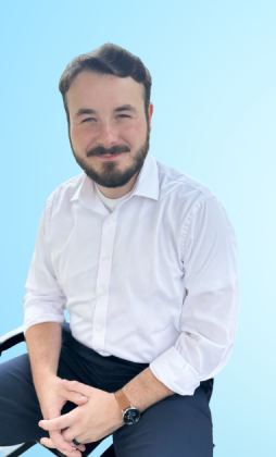 Portrait of a man with dark hair and beard, wearing a white shirt, sitting against a light blue background.