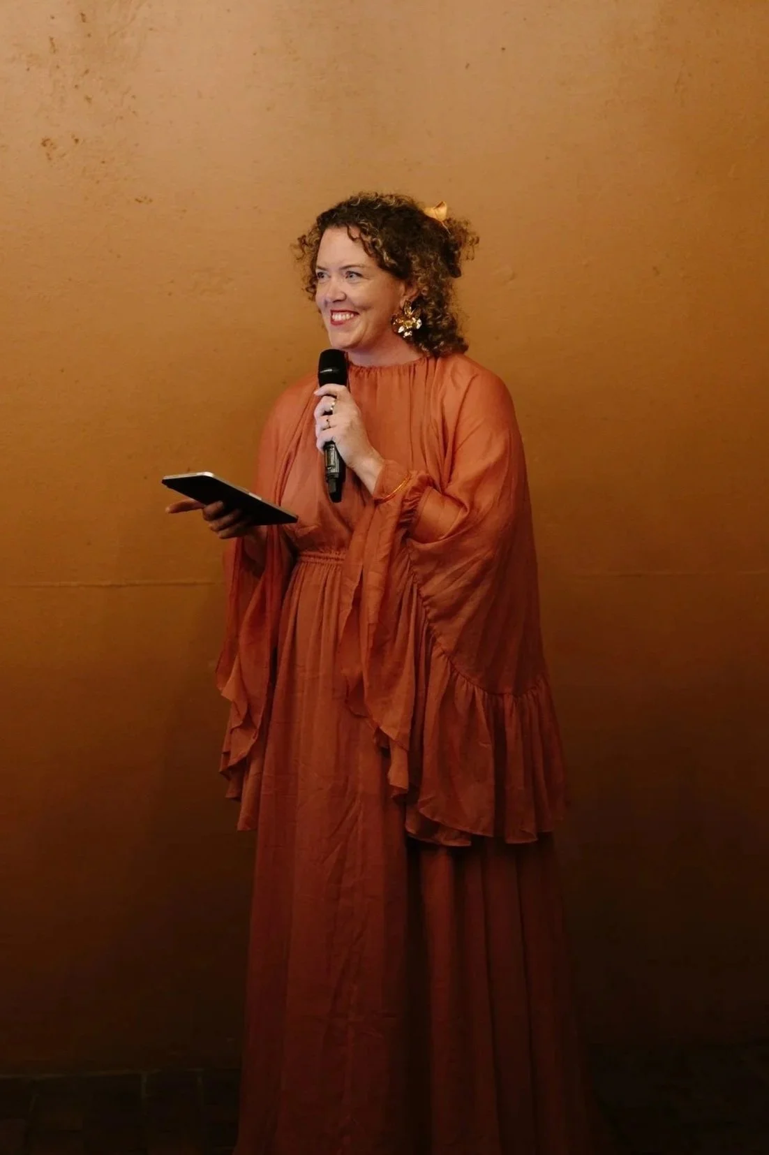Woman with curly hair wearing an orange dress, holding a microphone and a tablet, standing against a plain brown background.