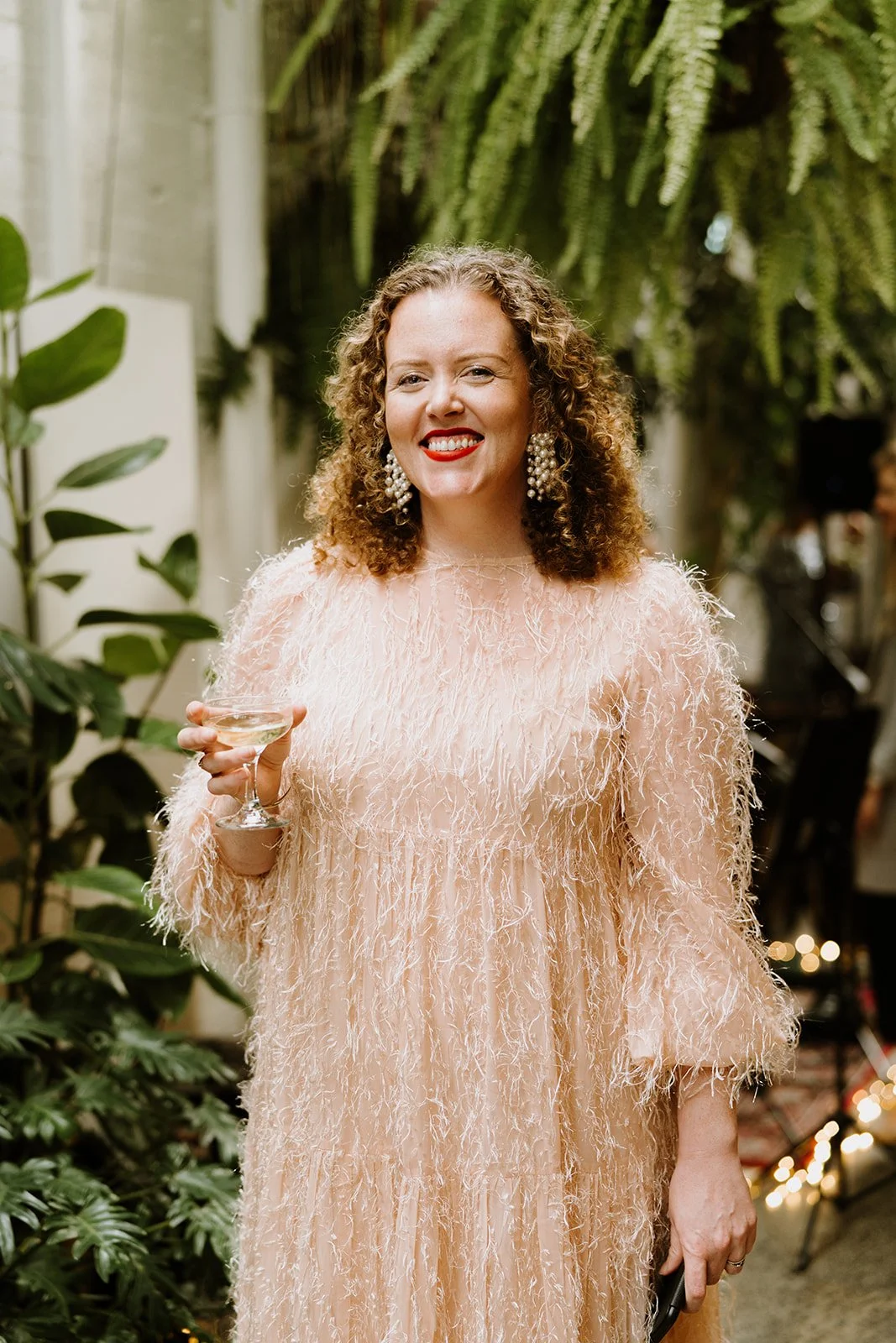 The Celebrant with curly hair wearing a peach-colored, feathered dress and large pearl earrings, smiling and holding a cocktail glass at an indoor event with lush greenery and hanging plants in the background.