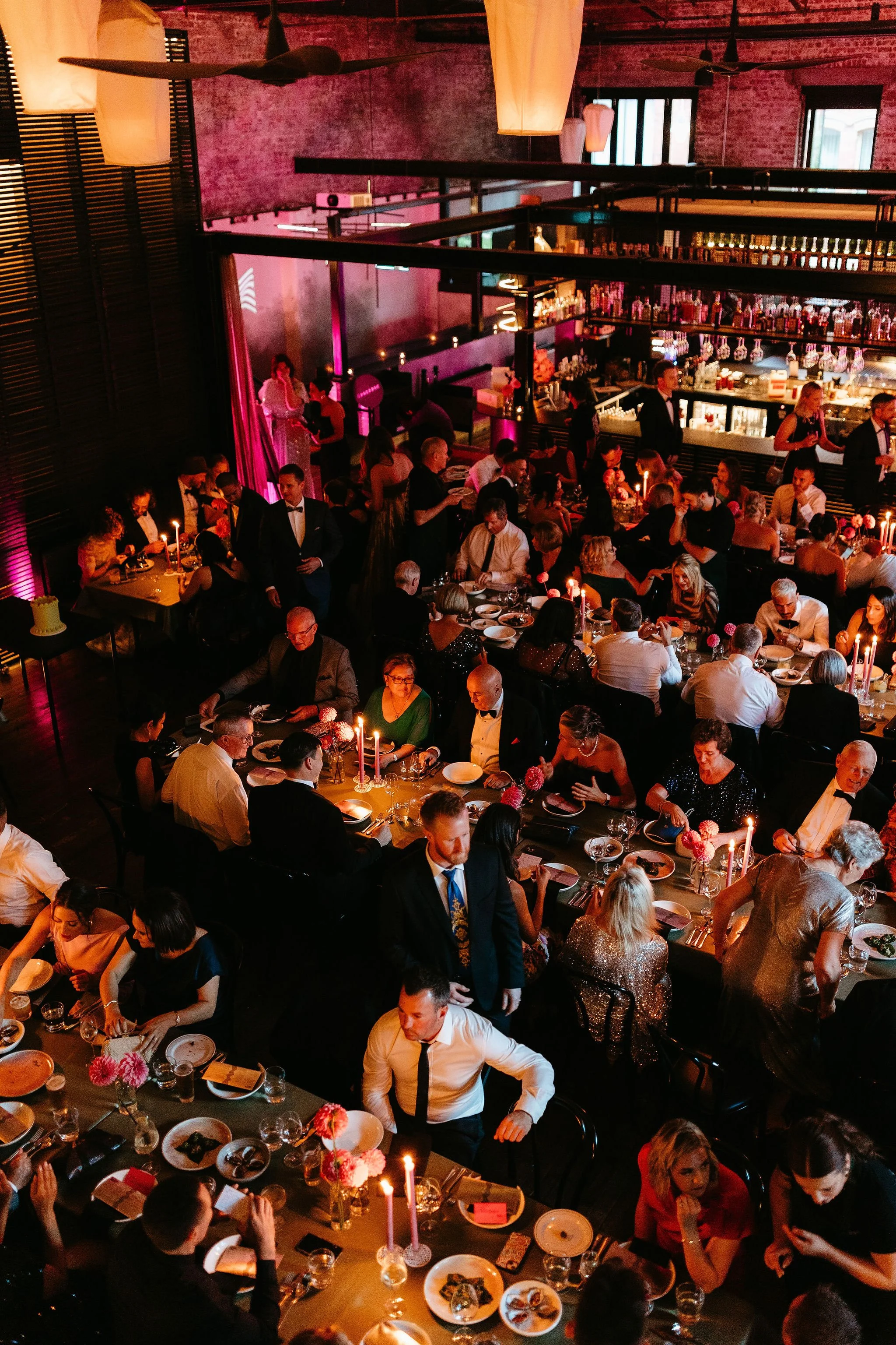 A crowded indoor dining event at a stylish restaurant or banquet hall with formal attire, candlelit tables, and a bar area in the background.