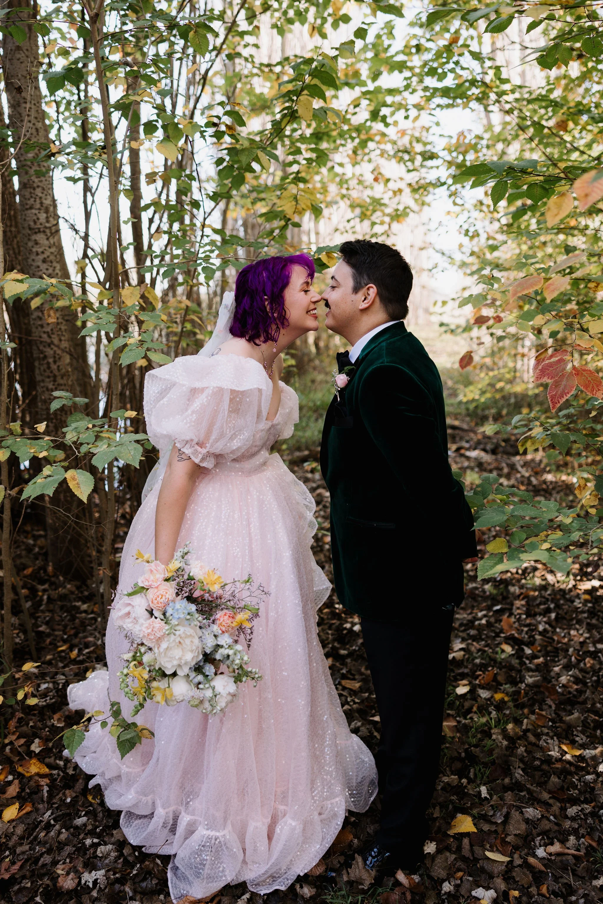 A bride with purple hair and a groom in a velvet green tuxedo leaning nose-to-nose in a forest setting, surrounded by autumn leaves, with the bride holding a bouquet of flowers.