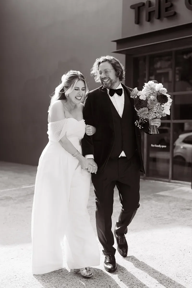 A black-and-white photo of a bride and groom walking together outdoors, smiling and holding hands. The bride is wearing a white off-shoulder wedding dress and the groom is dressed in a tuxedo with a bow tie, holding a bouquet of flowers.