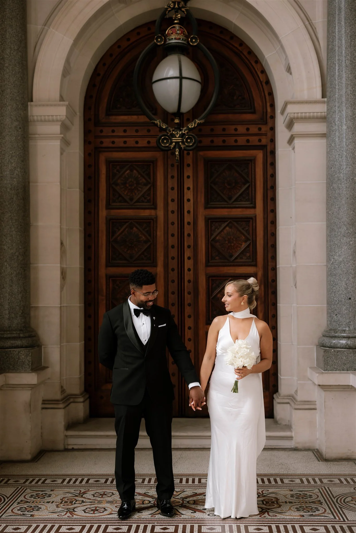 A bride and groom holding hands and smiling at each other in front of large wooden doors inside a historic building.