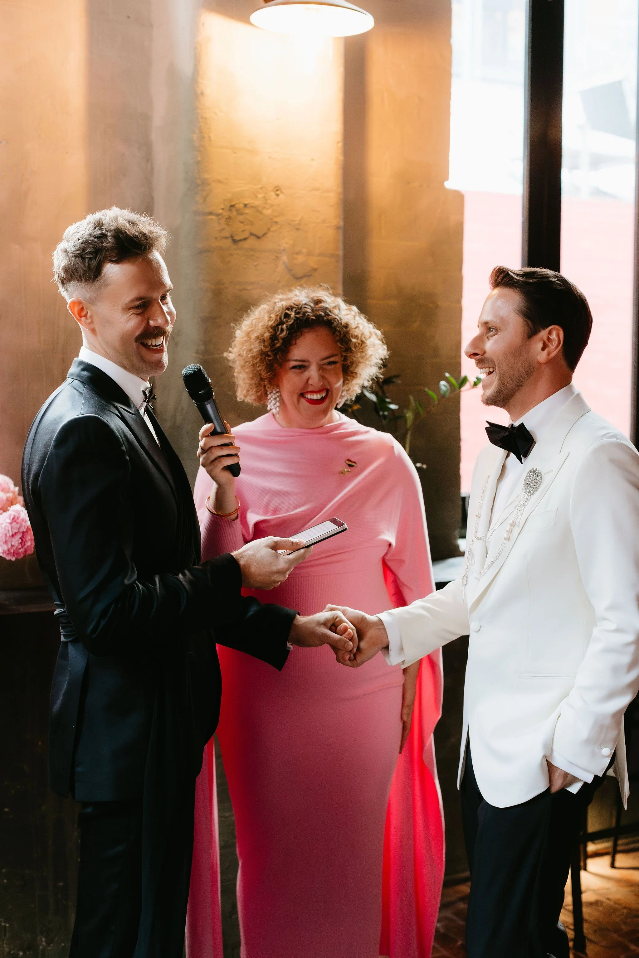 Two grooms holding hands and smiling at each other during their wedding ceremony, with an officiant standing between them, holding a microphone and reading vows.