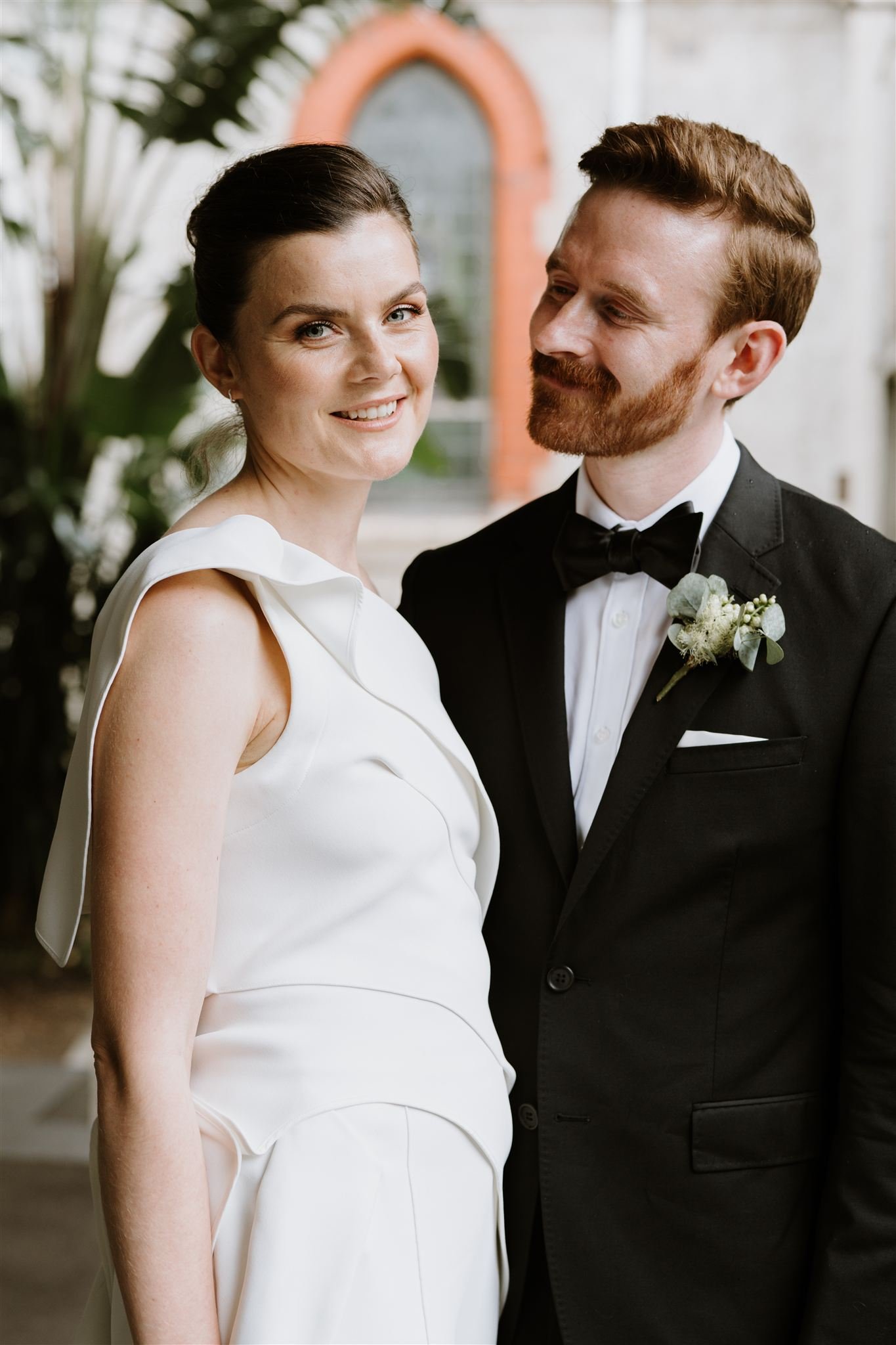 A bride in a white wedding jumpsuit smiling and a groom in a black tuxedo with a bow tie, standing outside near plants and a building with an arched window.