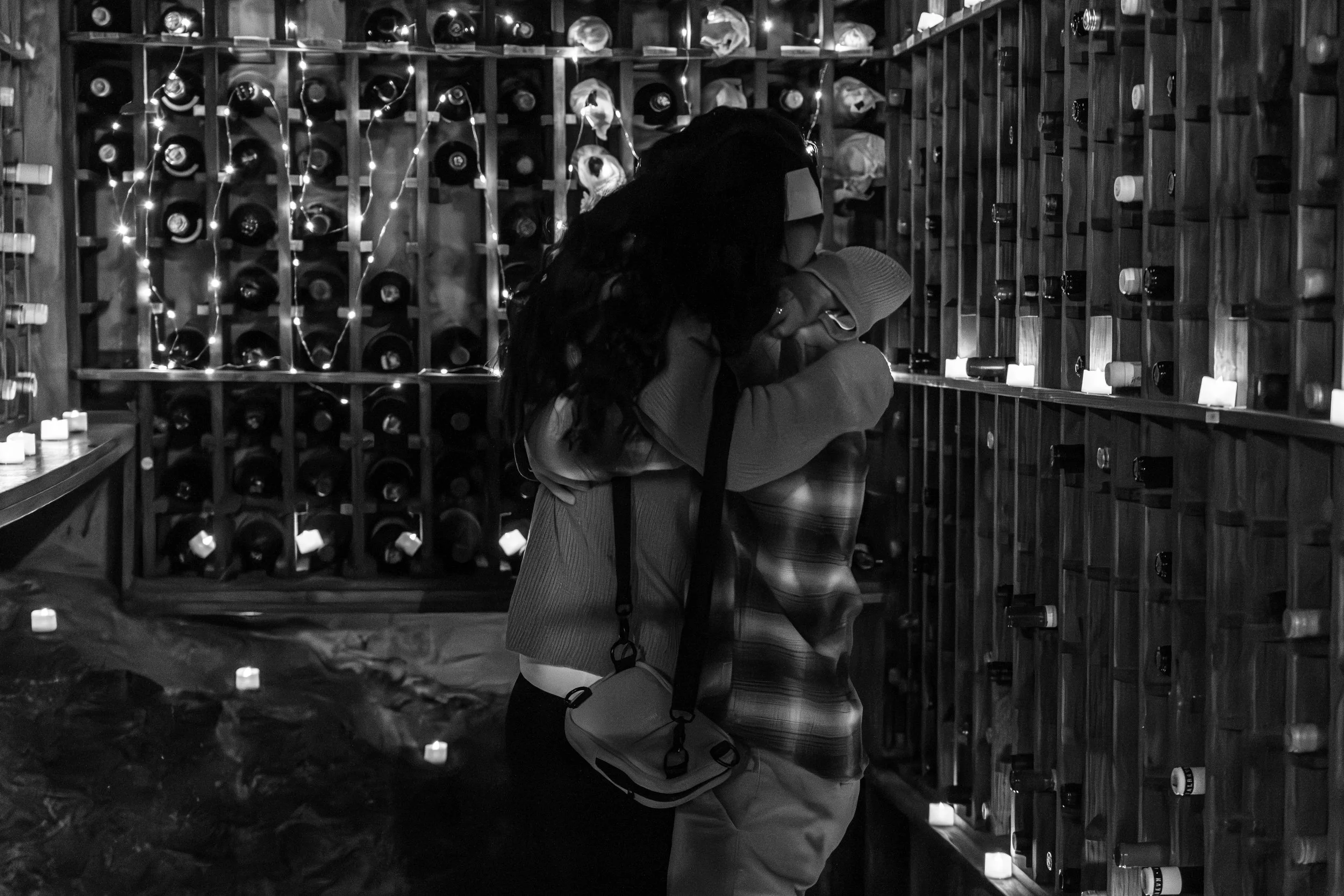 Proposal. Two people hugging in front of a wine rack with string lights and candles, in a wine cellar.