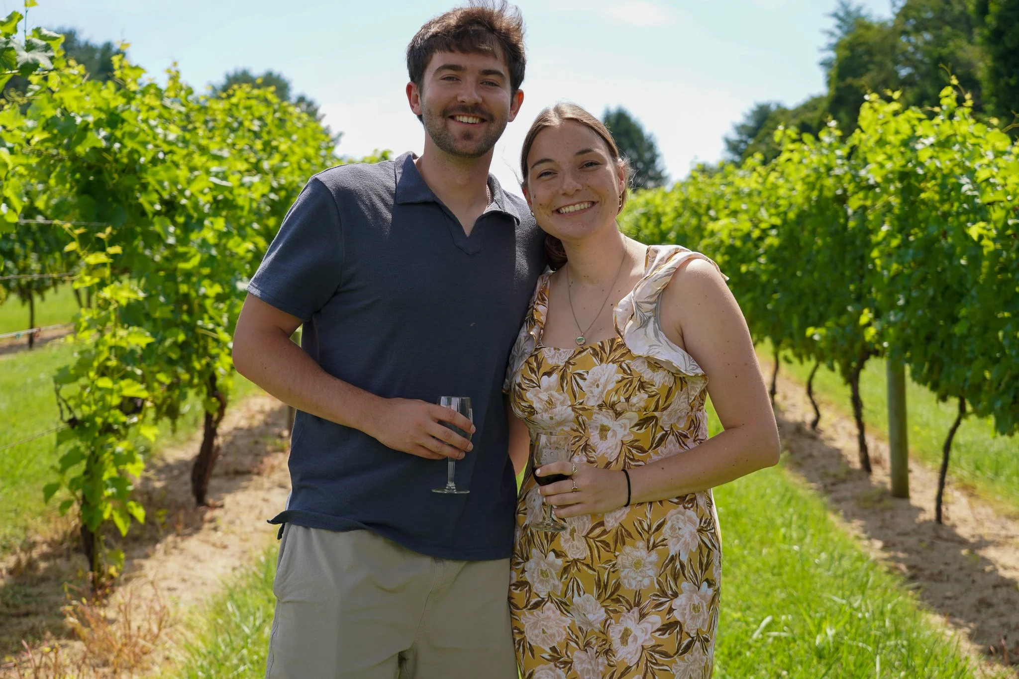 A smiling young man and woman stand together in a vineyard, each holding a glass of wine, during sunny weather. Maryland.