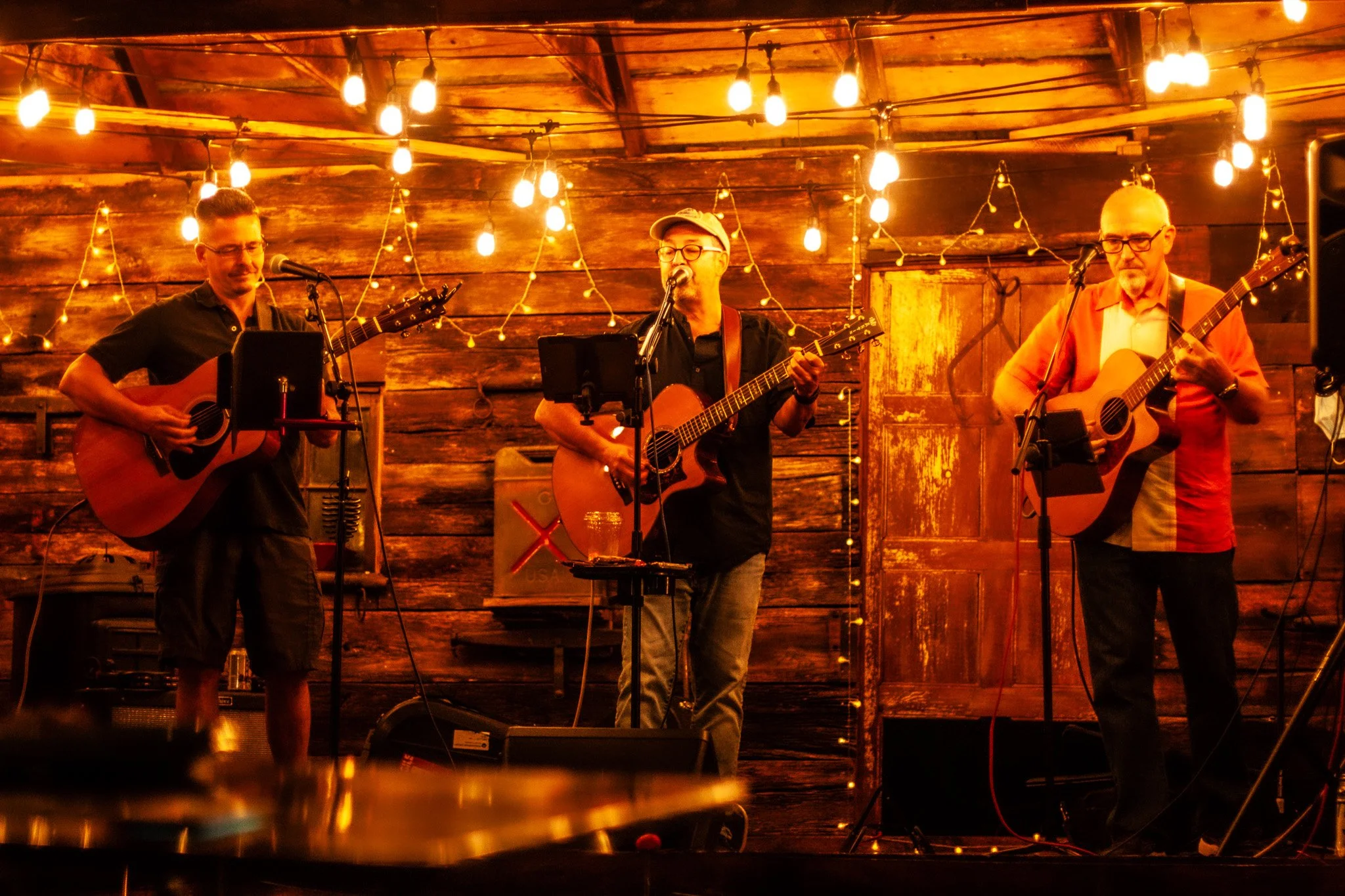 Three musicians playing acoustic guitars on a warmly lit wooden stage with string lights overhead.