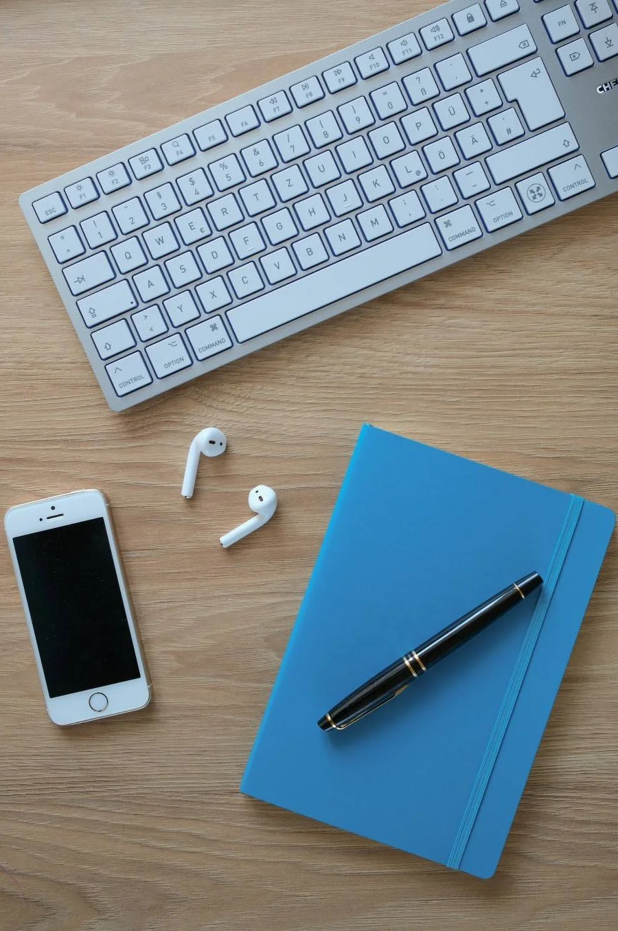 Computer-Tastatur, iPhone, kabellose Kopfhörer, blauer Notizbuch mit Kugelschreiber auf Holztisch.