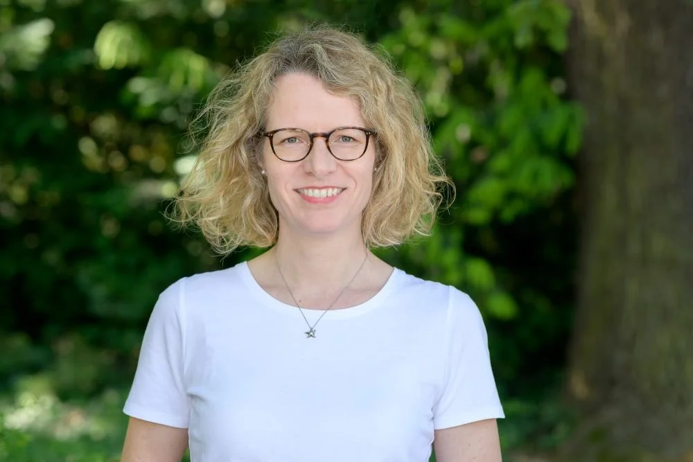 Eine lachende Frau mit lockigem blondem Haar, Brille und weißem T-Shirt vor einem grünen, bewaldeten Hintergrund.