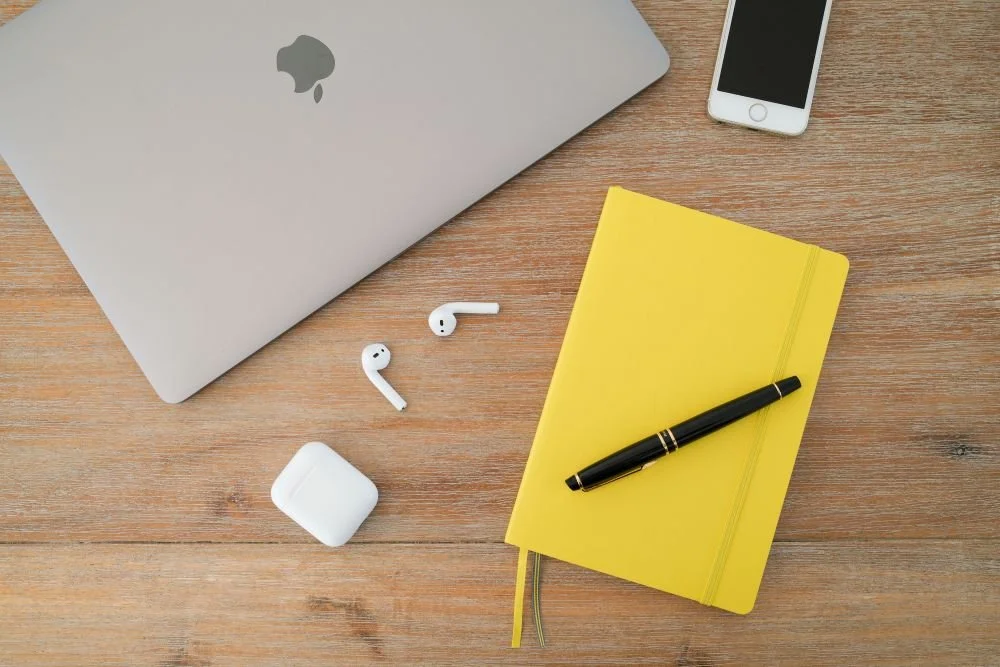 Ein MacBook, iPhone, AirPods, ein gelber Notizblock mit schwarzen Stift auf einem Holztisch.
