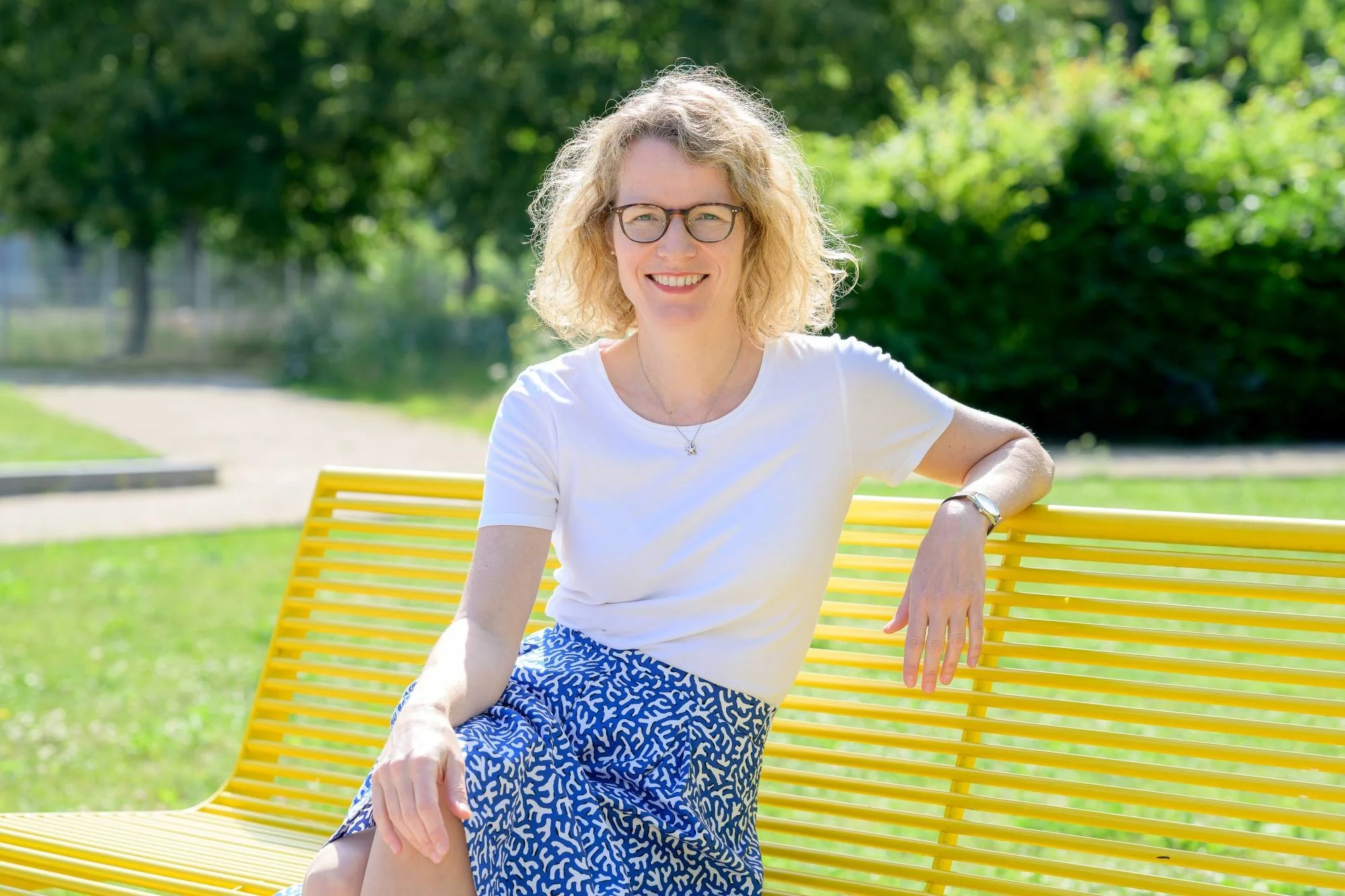 Eine lachende Frau mit Brille sitzt auf einer gelben Parkbank im sonnigen Park, umgeben von grünen Bäumen.