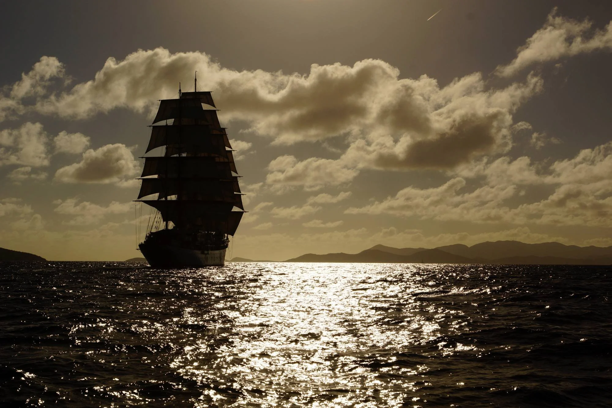 Segelschiff mit dunklen Segeln vor Sonnenuntergang, reflektierendes Wasser, Berge im Hintergrund, bewölkter Himmel.