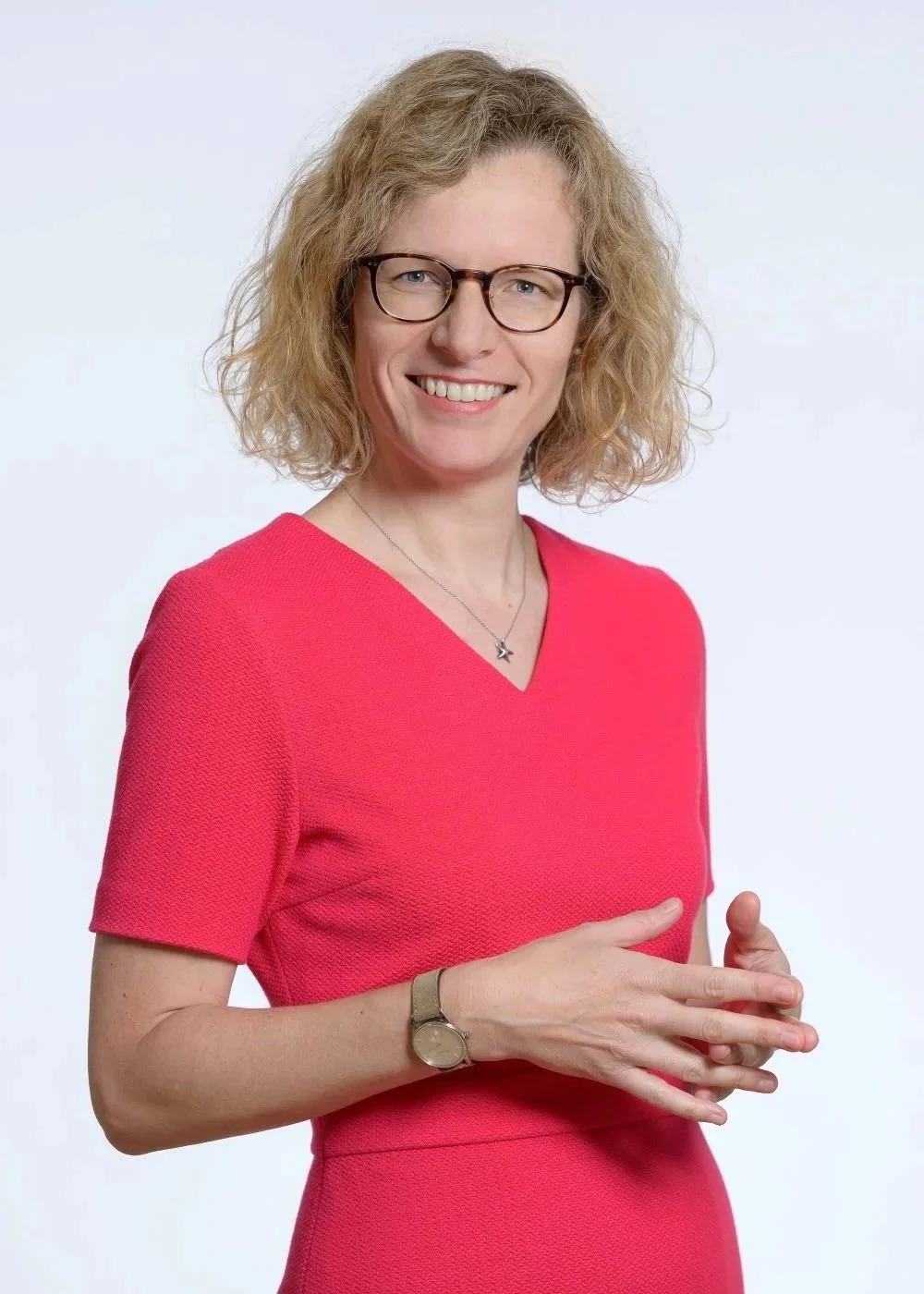 Frau mit Brille, Schulterlänge, lockigem blondem Haar, trägt eine rote V-Ausschnitt-Kleid und ein Armband, lächelt vor weißem Hintergrund.