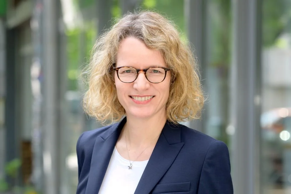 Porträt einer Frau mit lockigem, blondem Haar, Brille und dunklem Blazer, vor einem modernen Glasgebäude im Freien.