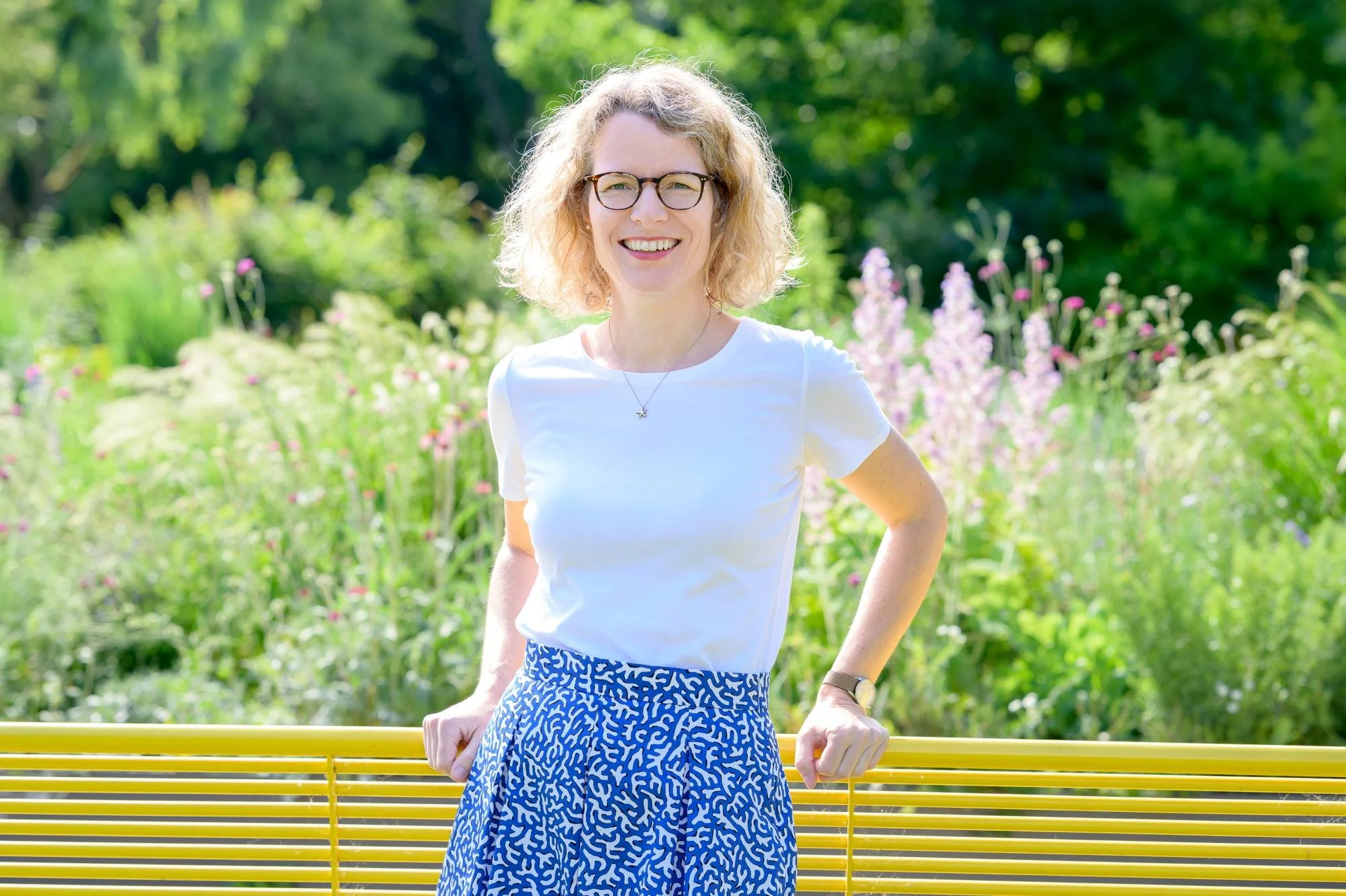 Lächelnde Frau mit Brille und blonden, lockigen Haaren steht im Park vor einer gelben Bank, im Hintergrund sind grüne Bäume und Blumen.