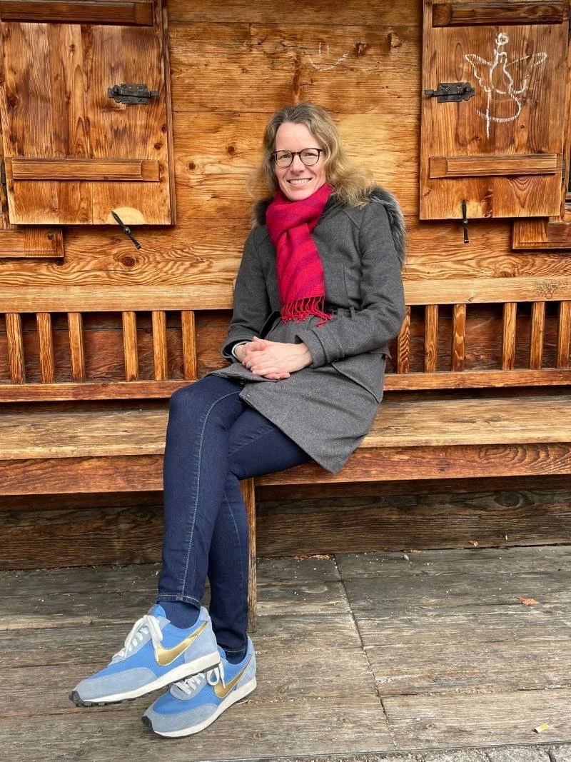 Frau sitzt auf einer Holzbank vor einer Holzwand mit Schiebetüren, trägt eine graue Jacke, eine rote Schal, blaue Jeans und Sneakers, lächelt und trägt Brille.