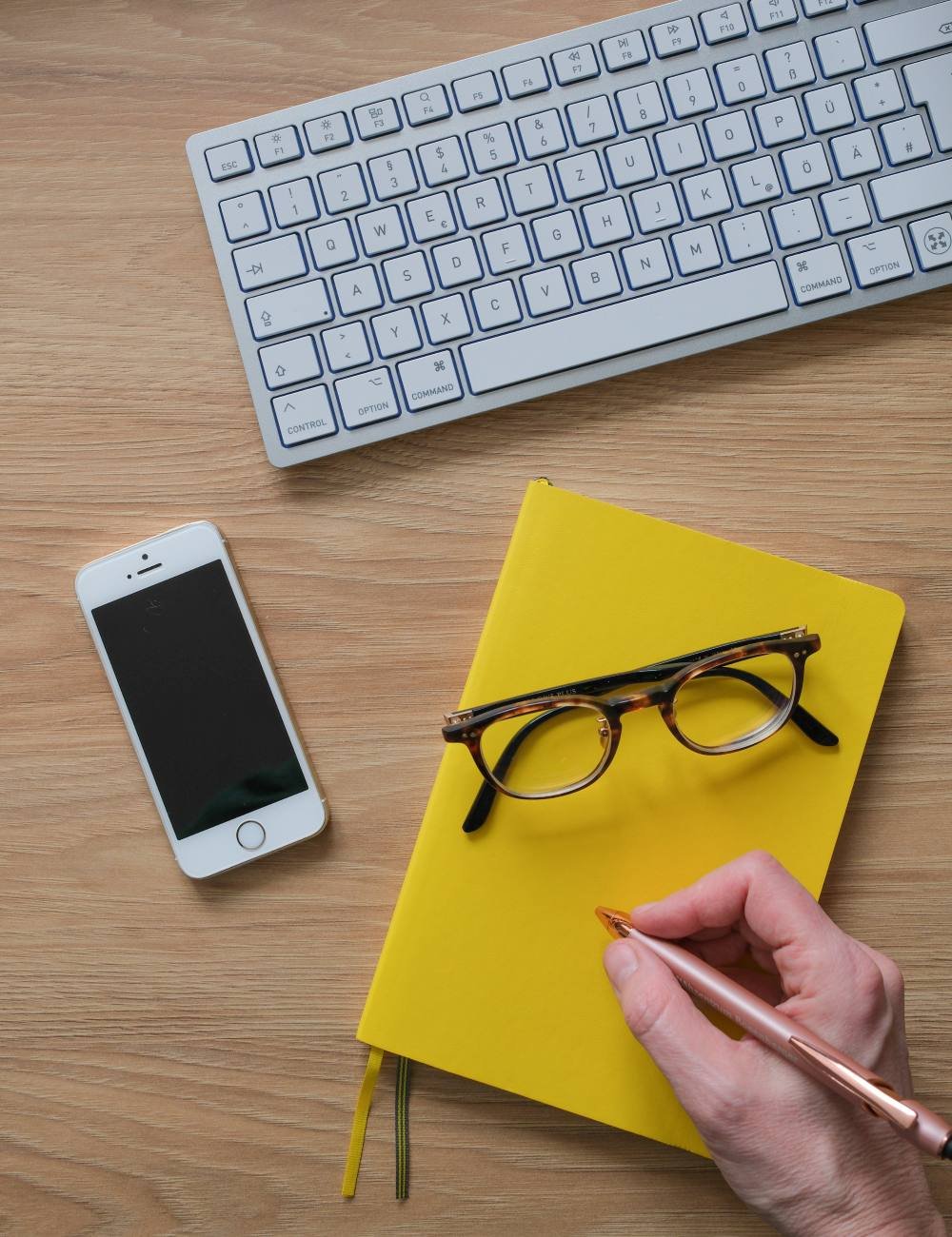 Ein Schreibtisch mit einem weißen Tastatur, einem Smartphone, einer gelben Notizbuch, einer Brille und einer Hand, die einen Stift hält.