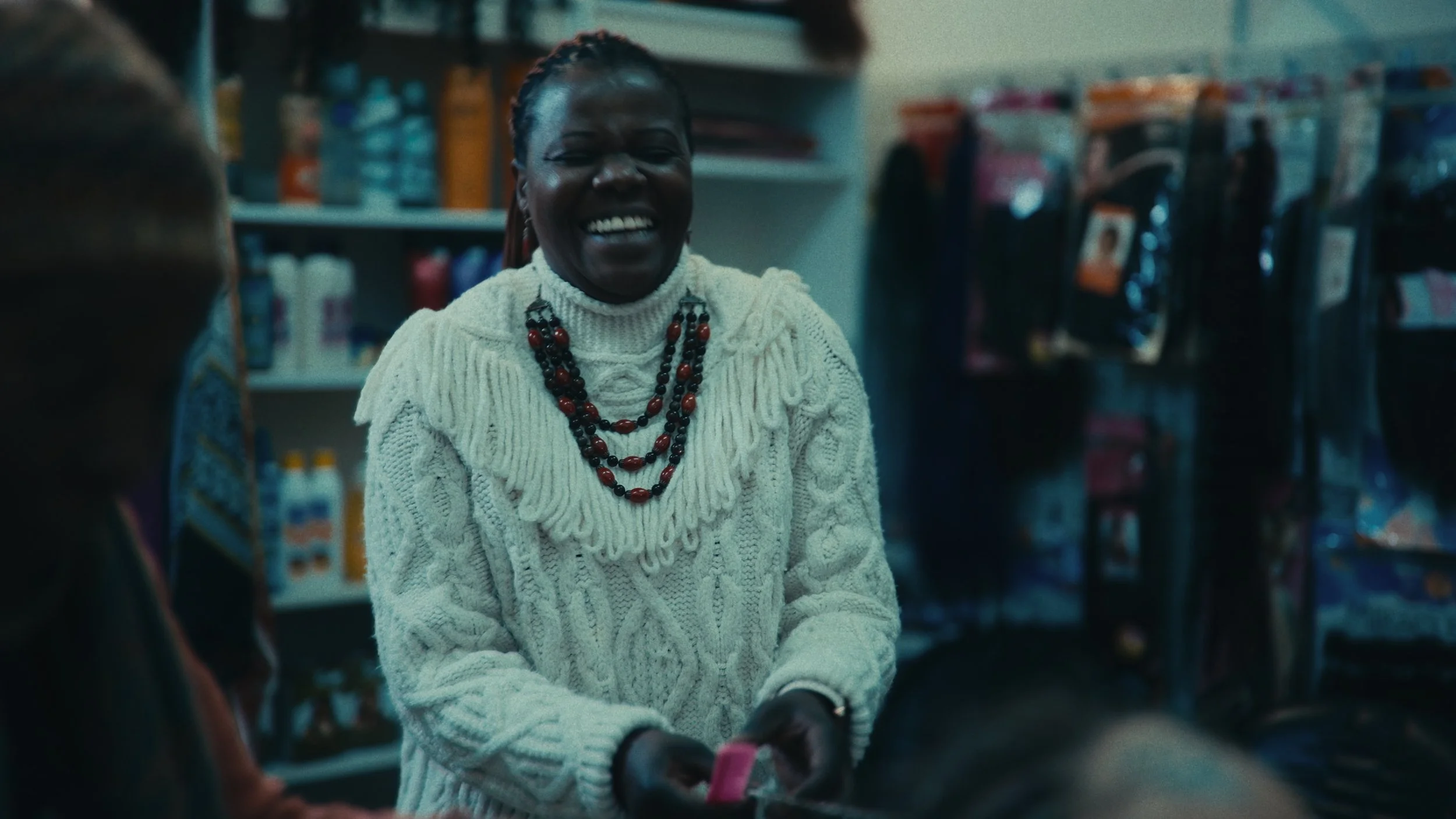 Una donna sorridente in un negozio circondata da altri clienti, indossa un maglione bianco lavorato a maglia e collane di perle nere e rosse.