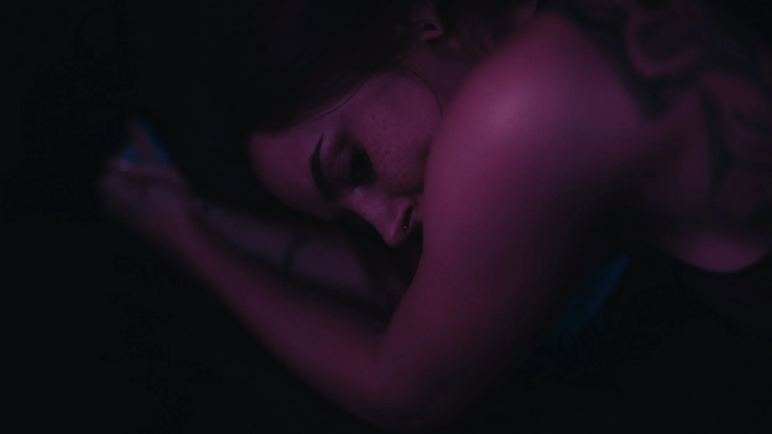 Ragazza con capelli scuri e tatuaggio, che dorme su un letto in una stanza con luci soffuse rosa e viola.