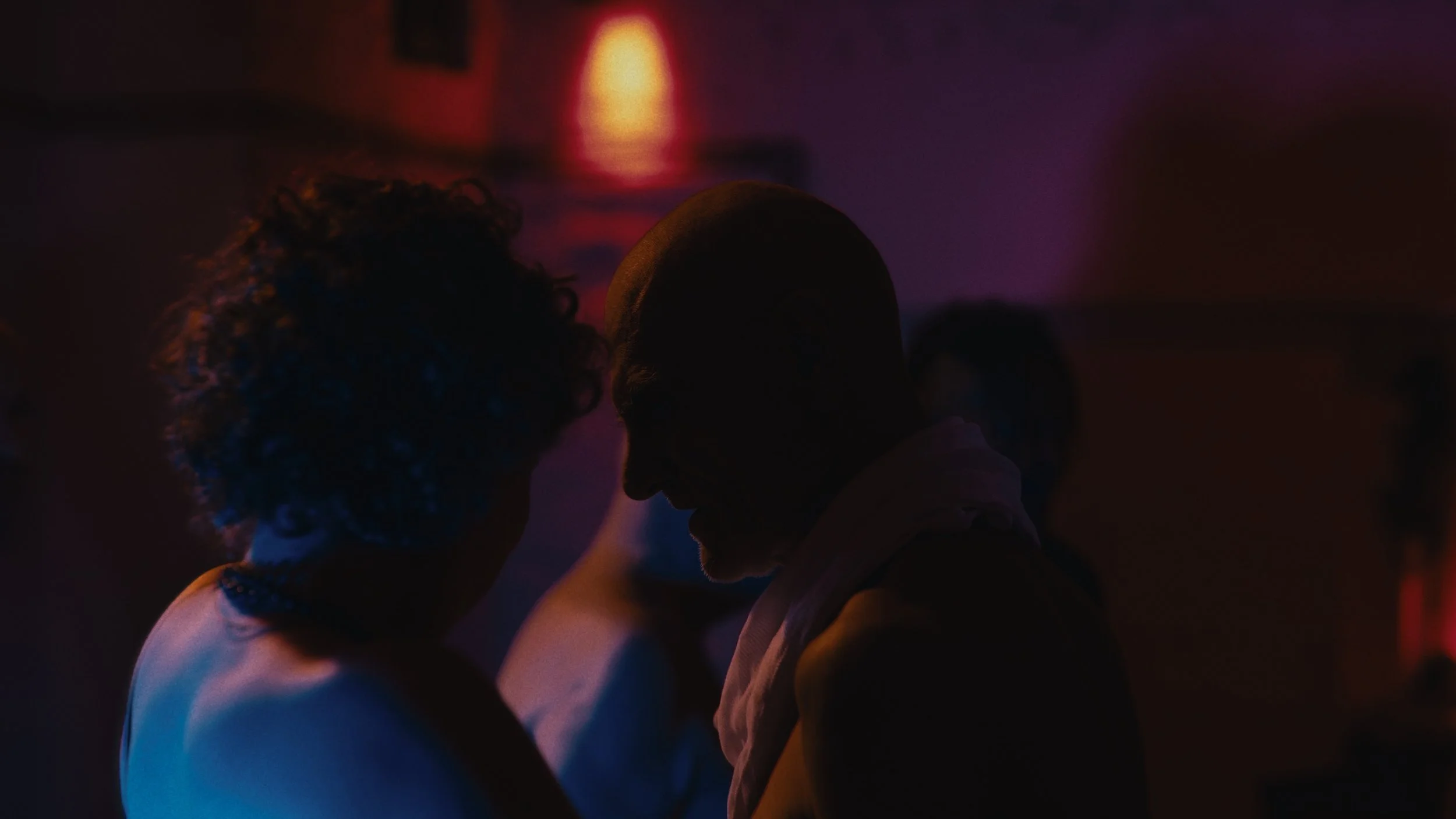 Due persone in un ambiente buio con luci colorate, si trovano molto vicine e si guardano con intimità.