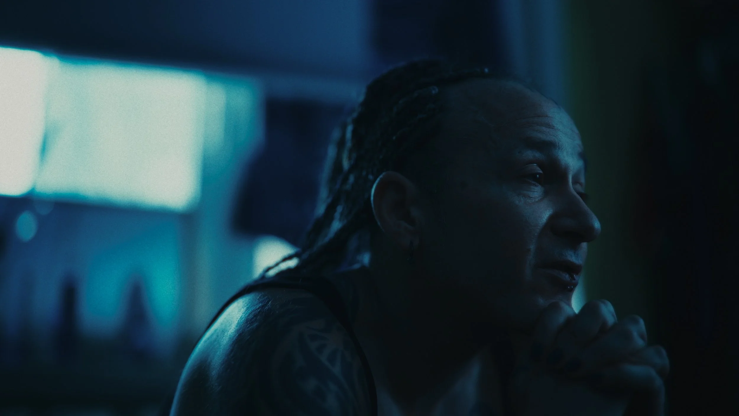 Persona con capelli raccolti e tatuaggi, in vibe notturno, guarda pensierosa in ambientazione dark e futuristica.