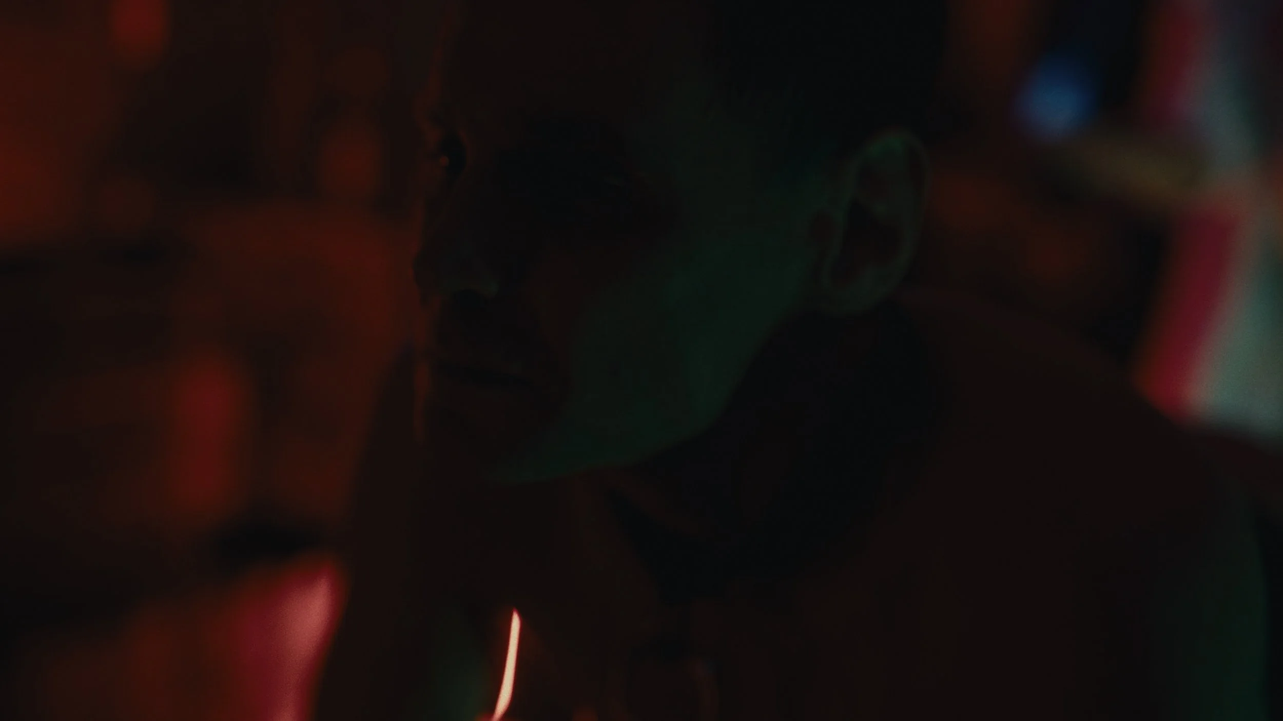 Immagine scura di un uomo con orecchiali, con luce rossa e verde che illuminano parzialmente il suo volto, in un ambiente forse notturno o in un luogo con illuminazione soffusa.