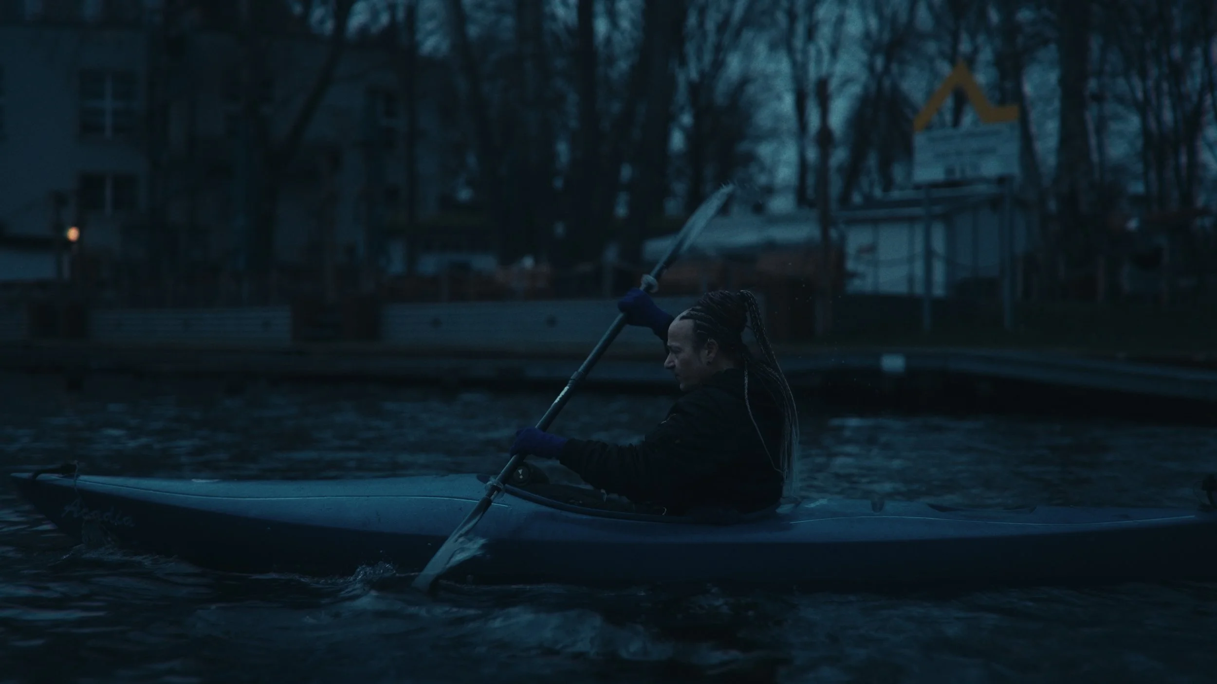 Persona che paga una pagaia in una barca in acque calme al crepuscolo con edifici e alberi sullo sfondo.