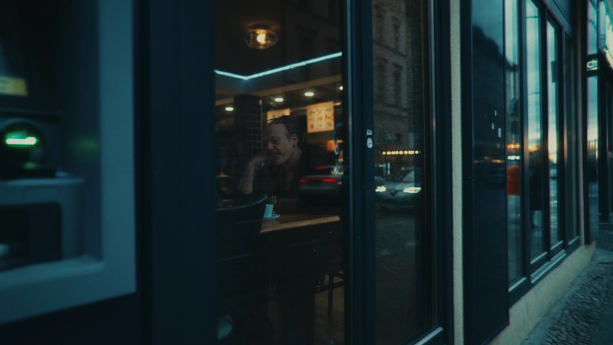 Cliente seduto al tavolo di un ristorante vista attraverso la finestra, con luci soffuse e un ambiente interno accogliente, all'esterno una strada con luci serali.