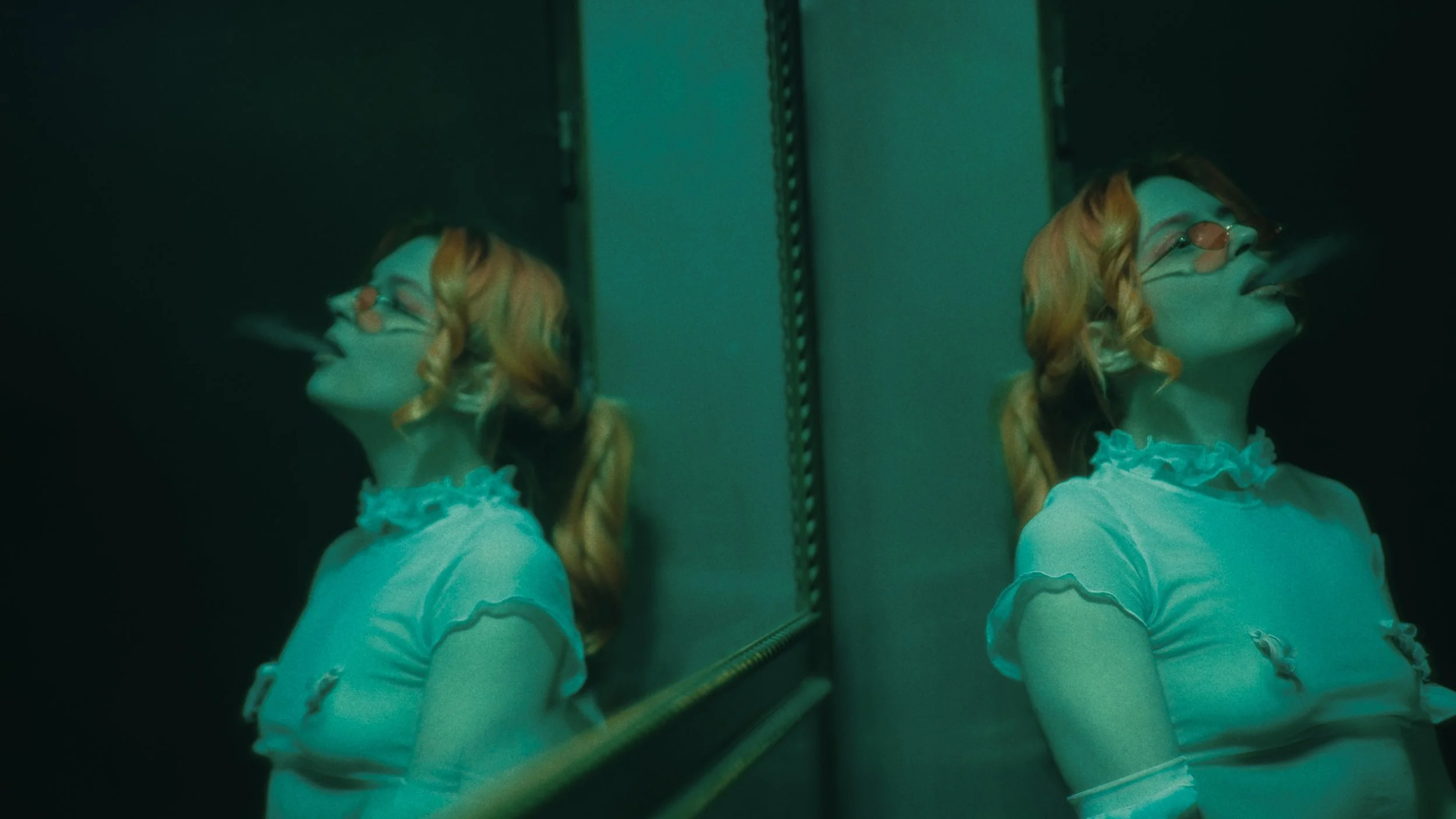 Una donna con capelli riccioli rossi, indossa occhiali arancioni e una maglietta rosa con dettaglio di volant, si fuma una sigaretta davanti a uno specchio.