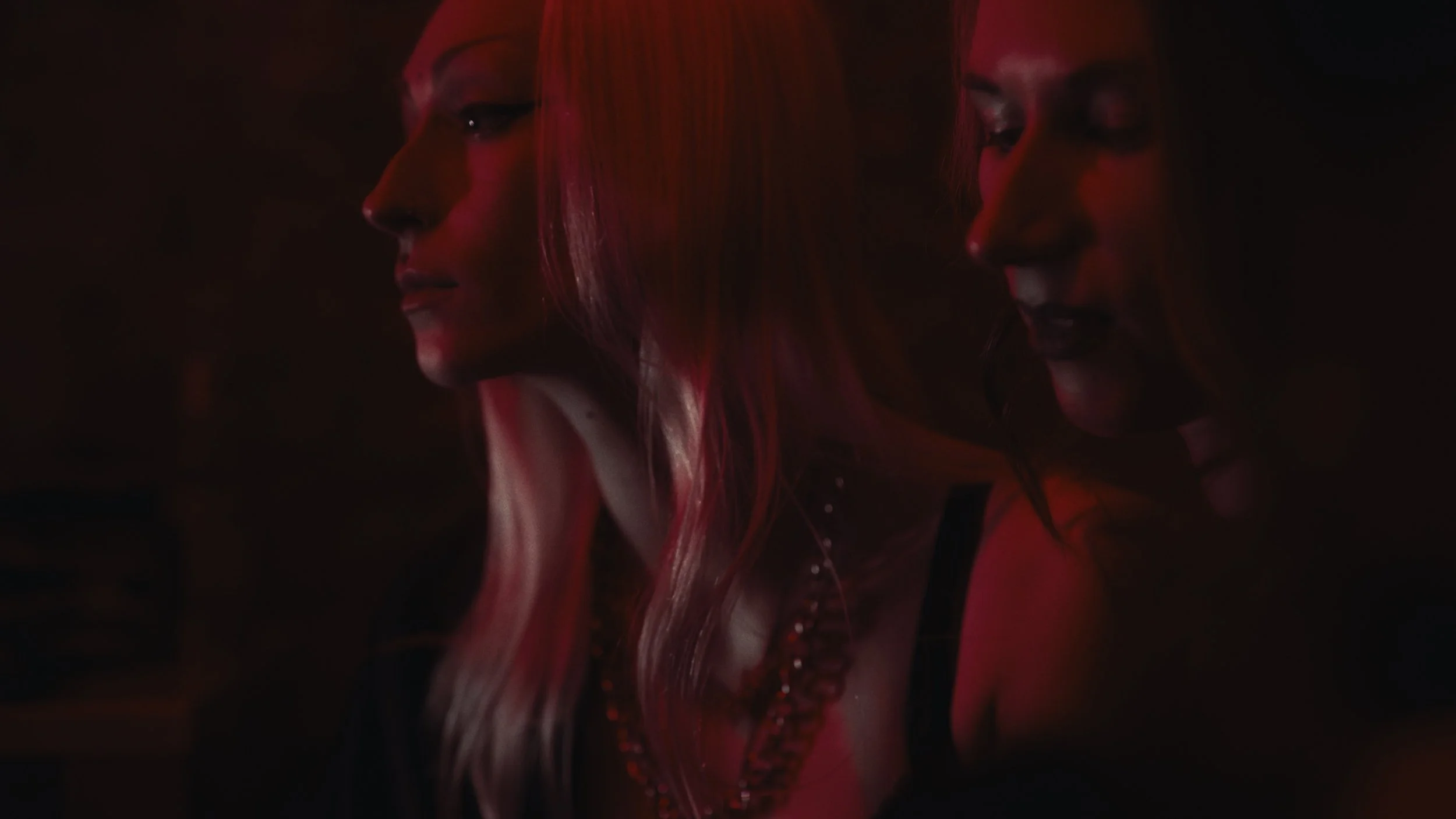 Due donne con capelli lunghi, uno con capelli biondi e l'altro con capelli rossi, in un ambiente con luce soffusa e colori caldi, che sembrano conversare o condividere un momento.