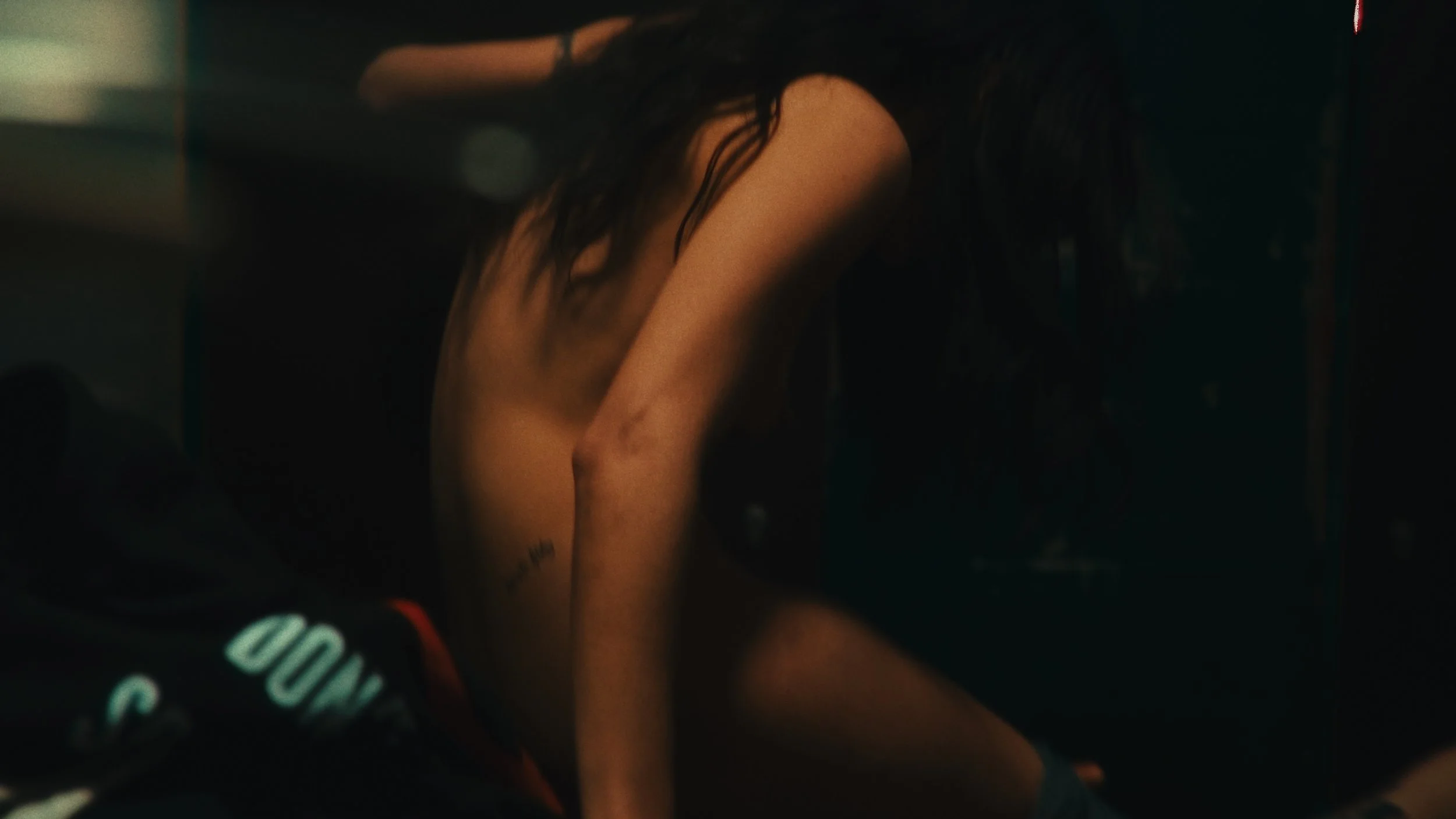 Immagine scura di una donna nuda con capelli ricci, seduta con le gambe piegate, in un'ambientazione poco illuminata.