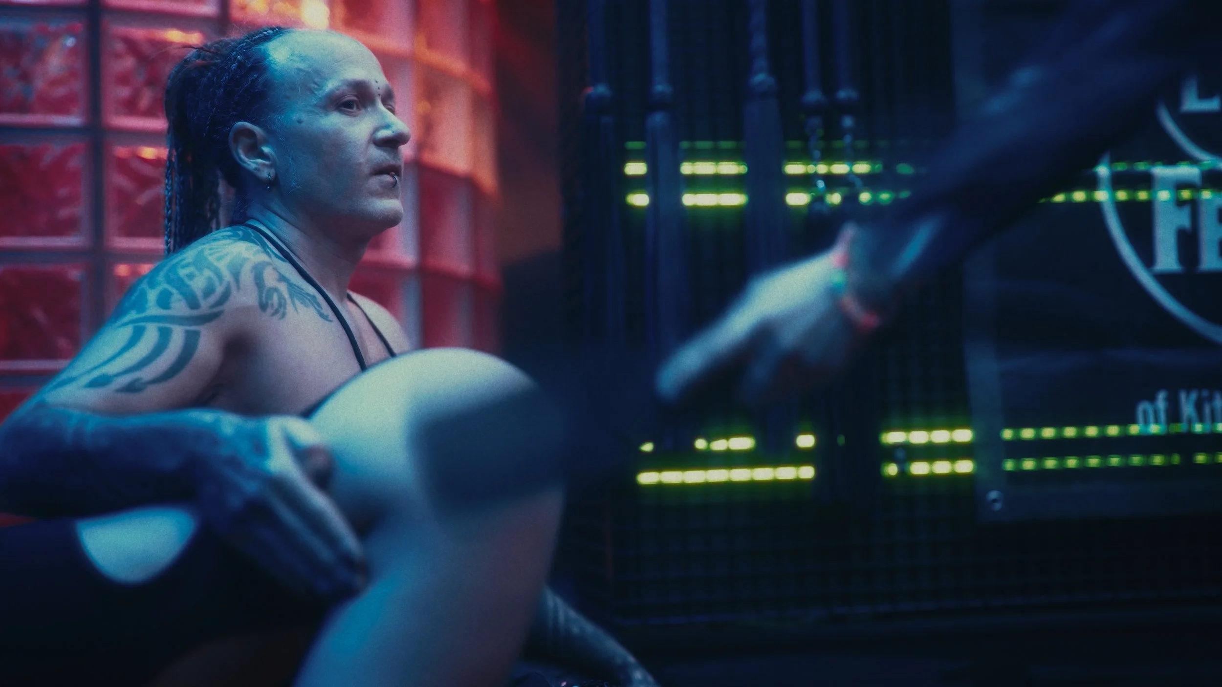 Una donna con tatuaggi sui bracci e sulle spalle, con i capelli in treccia, si trova in un ambiente con luci al neon di colore blu e rosso, con un calcio e un'asta di football americano, in una scena che sembra essere un allenamento o un'ambientazion