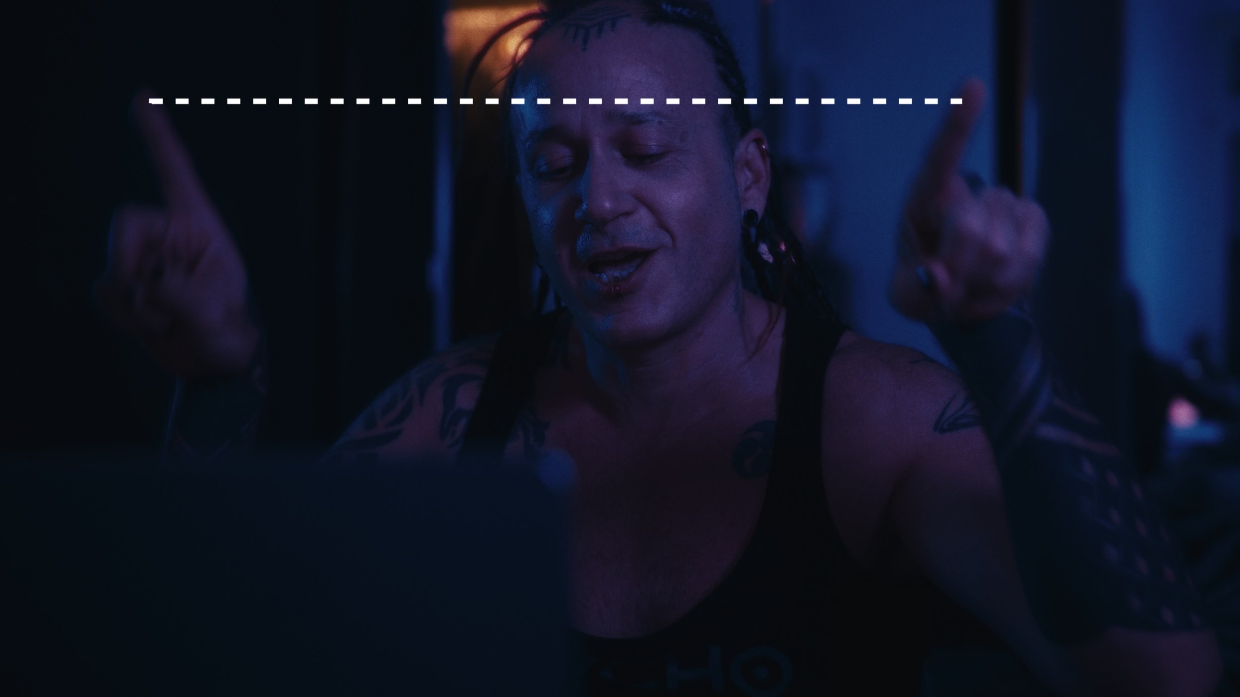 Uomo con tatuaggi, capelli a treccia, in abbigliamento nero, che sorride mentre guarda uno schermo in un ambiente scuro, illuminato da luci blu e viola.