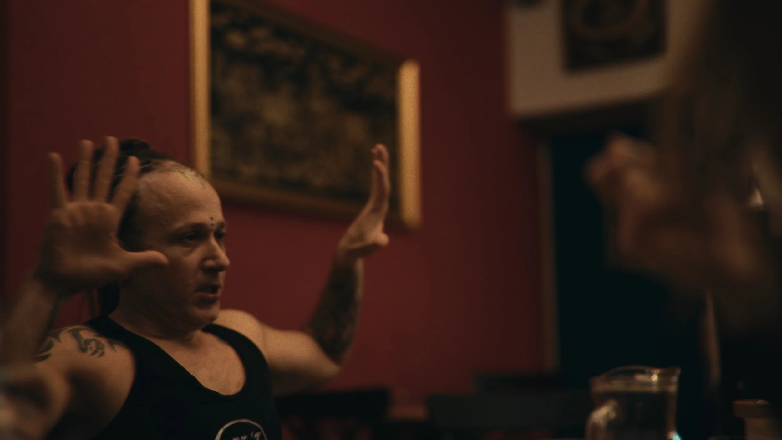 Persona con capelli biondi, tatuaggi, e una maglietta nera, fa un gesto con le mani in un locale con pareti rosse e quadri alle pareti.