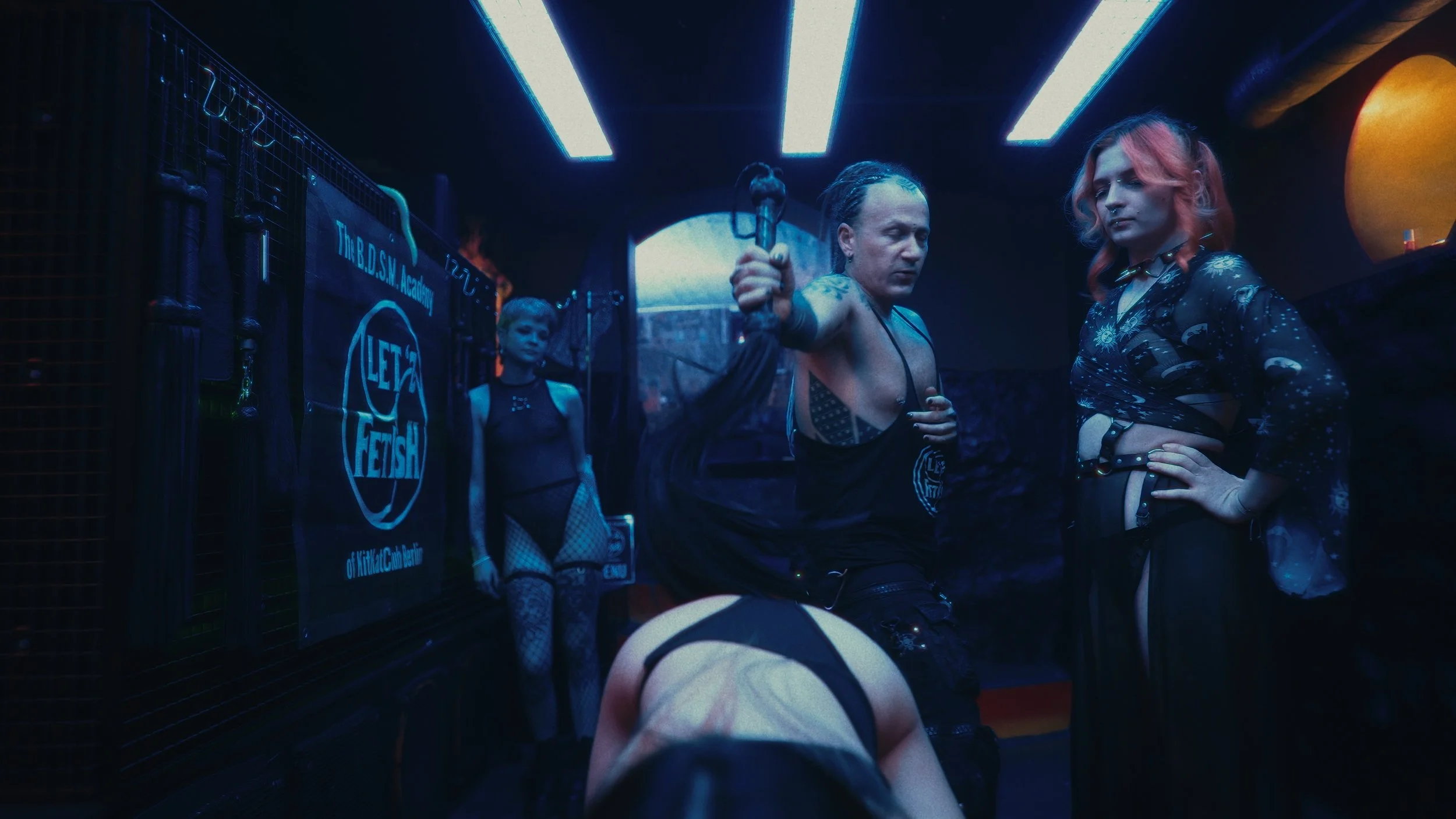 Scene in una sala buia con neon blu, persone vestite in abiti fetish, tra cui una donna con un cappio in mano e altre in pose provocative, con un banner che dice 'Let Fetish'.