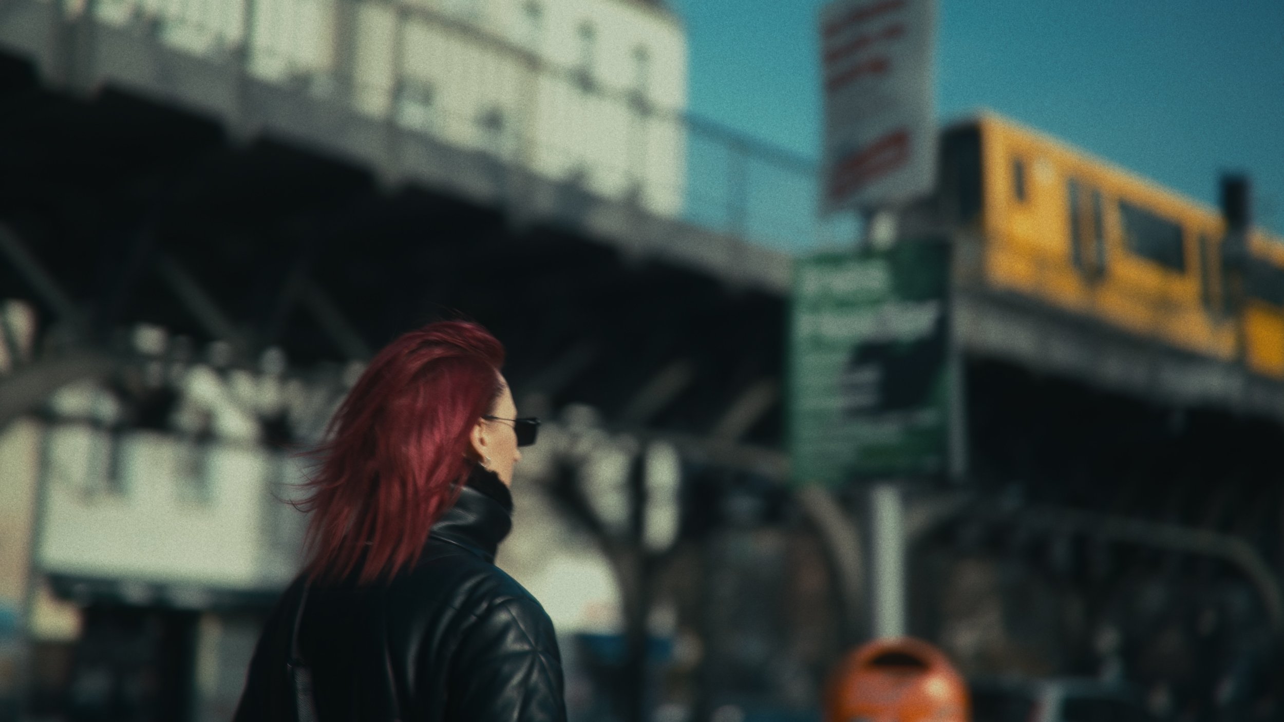 Una donna con capelli rossi e giacca di pelle guarda verso destra, in un ambiente urbano con ferrovie sopraelevate e un treno giallo in lontananza