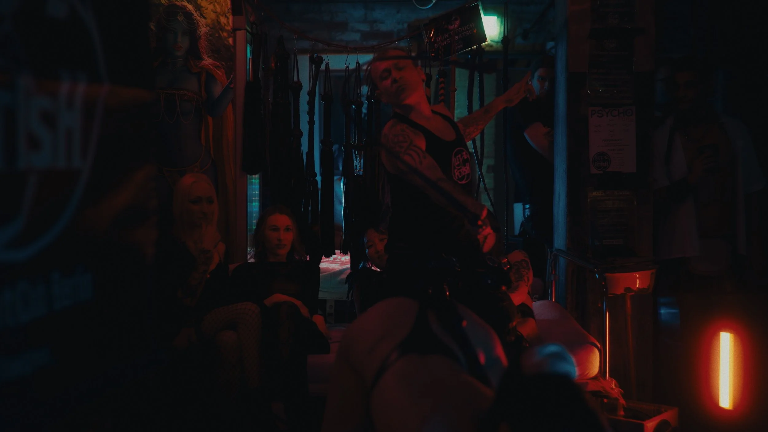 Una stanza oscura illuminata da luci rosse e blu, con persone sedute e in piedi. Al centro, una donna con tatuaggi che indossa abiti provocanti e sta seduta su una persona sulla sedia, circondata da accessori come fruste e manette.